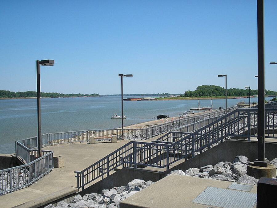A waterfront promenade with a concrete walkway, metal railings, and a series of street lamps.