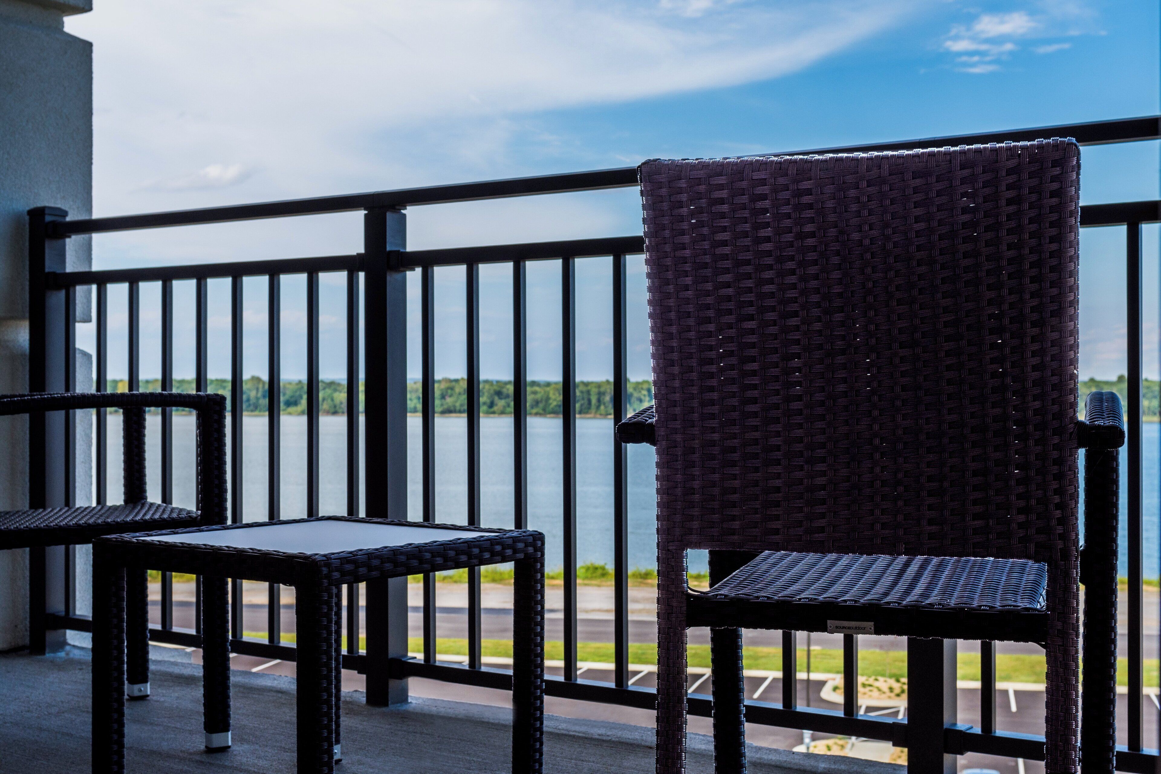 A balcony with a wicker chair and a table, overlooking a body of water and greenery.