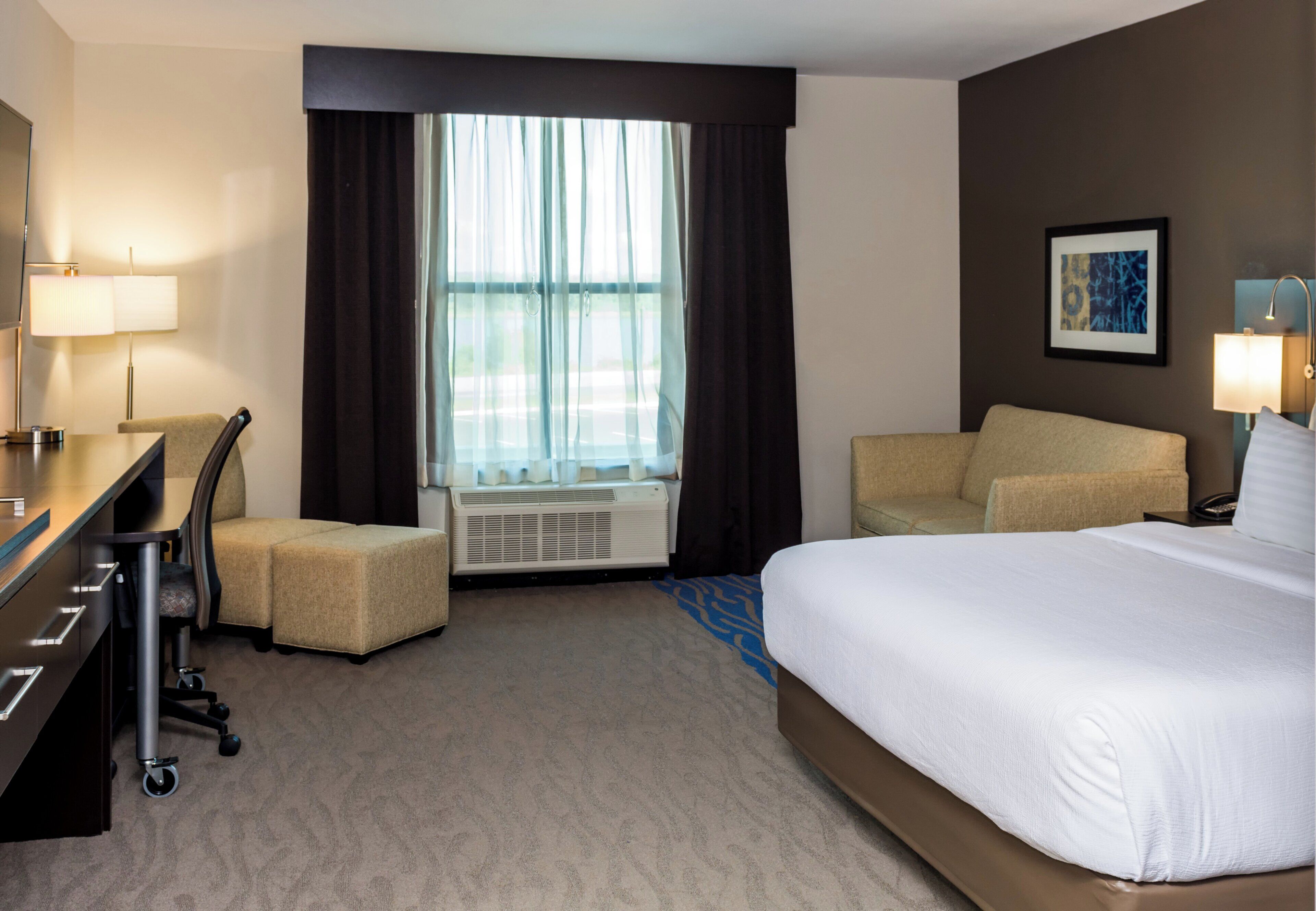 A hotel room with a bed, desk, chair, air conditioning unit, and a window with curtains.
