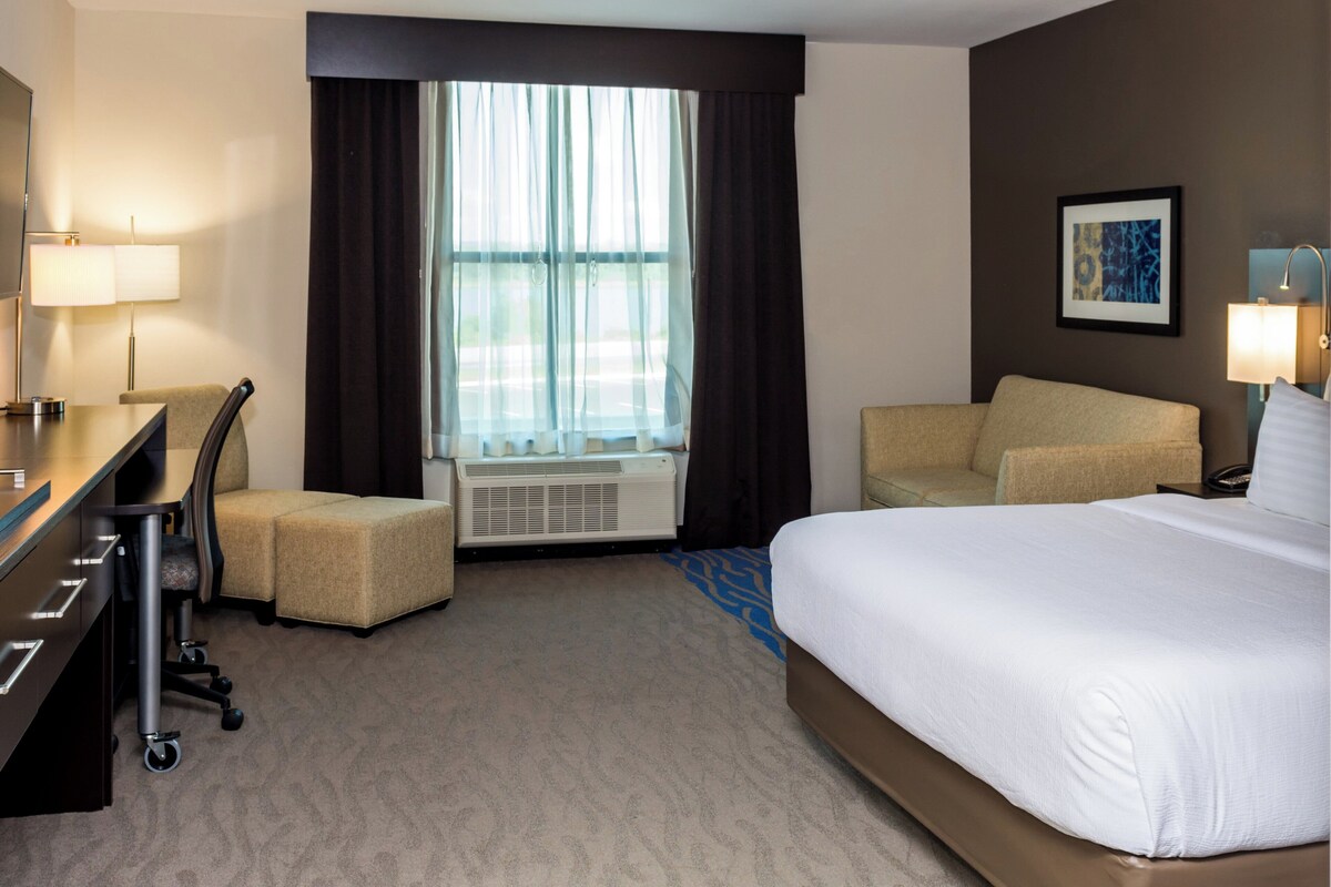 A hotel room with a bed, desk, chair, air conditioning unit, and a window with curtains.