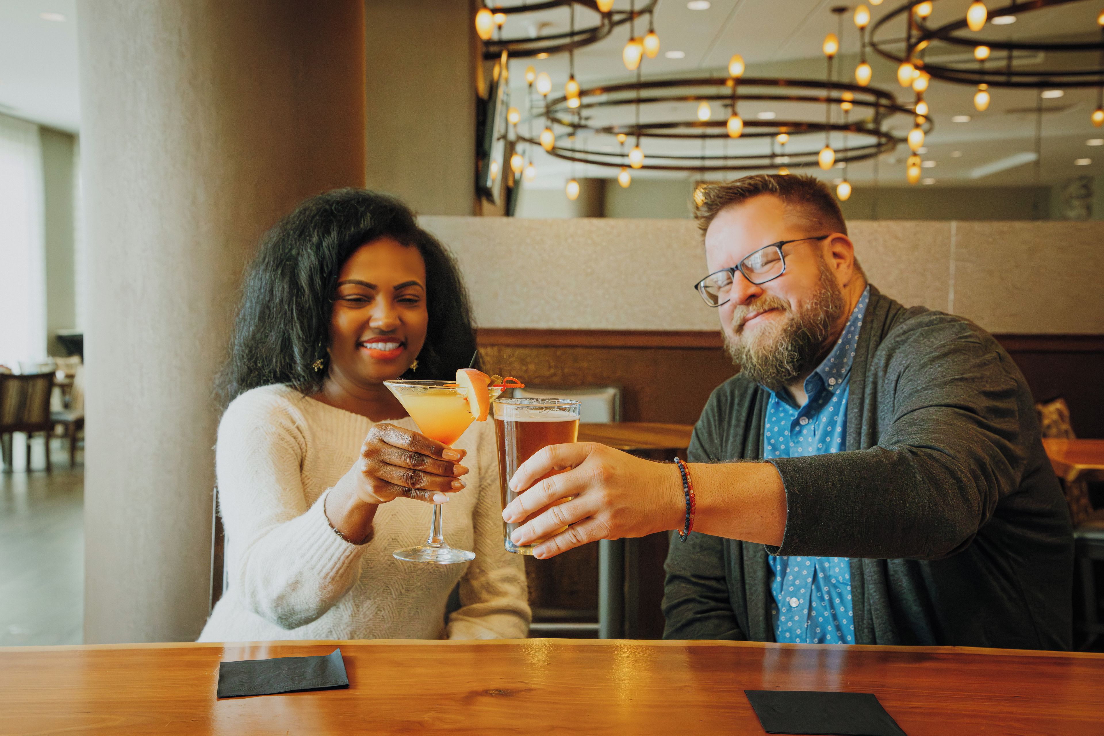Two people toasting with drinks at a bar with a modern interior and a visible ceiling light fixture.