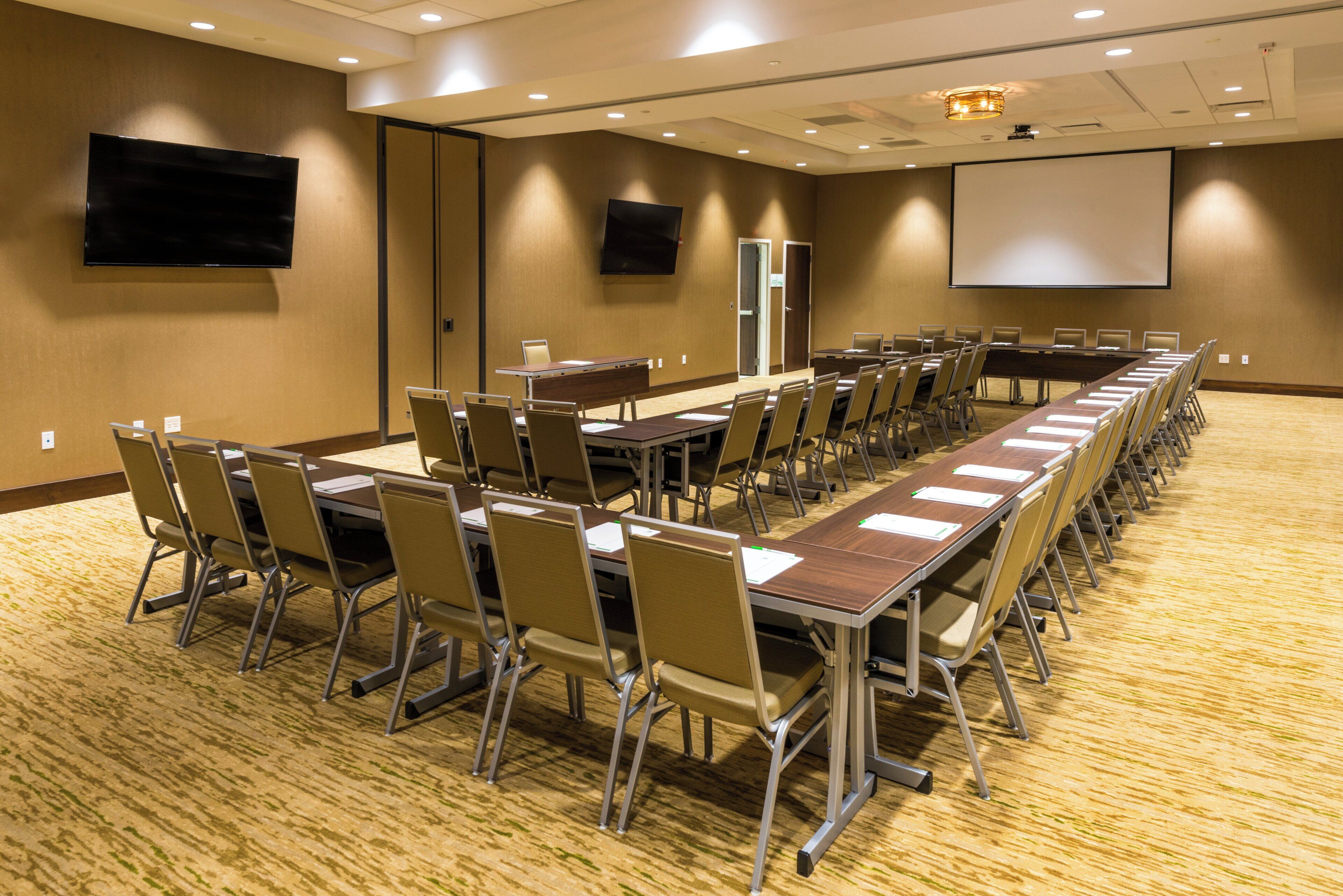 A conference room with long tables, chairs, a projector screen, and a television.