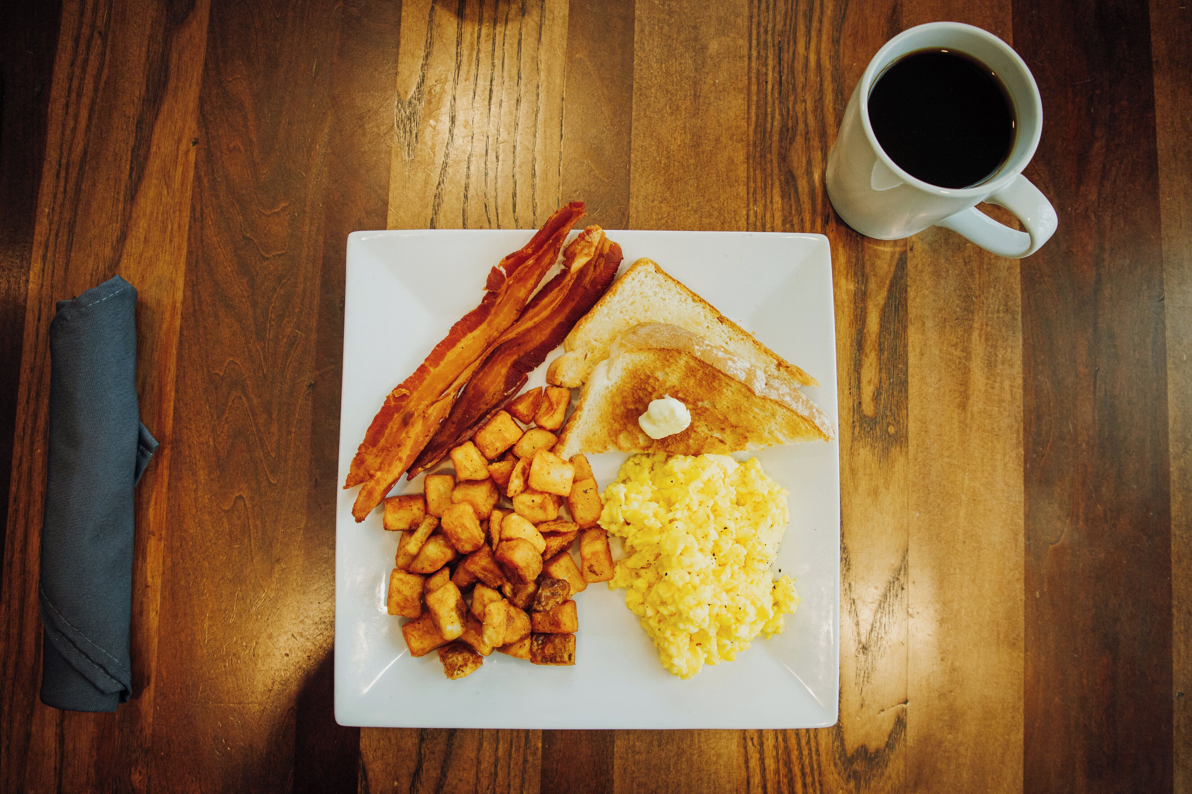 Un plato de desayuno con tocino, tostadas, huevos revueltos y patatas ralladas sobre una mesa de madera.