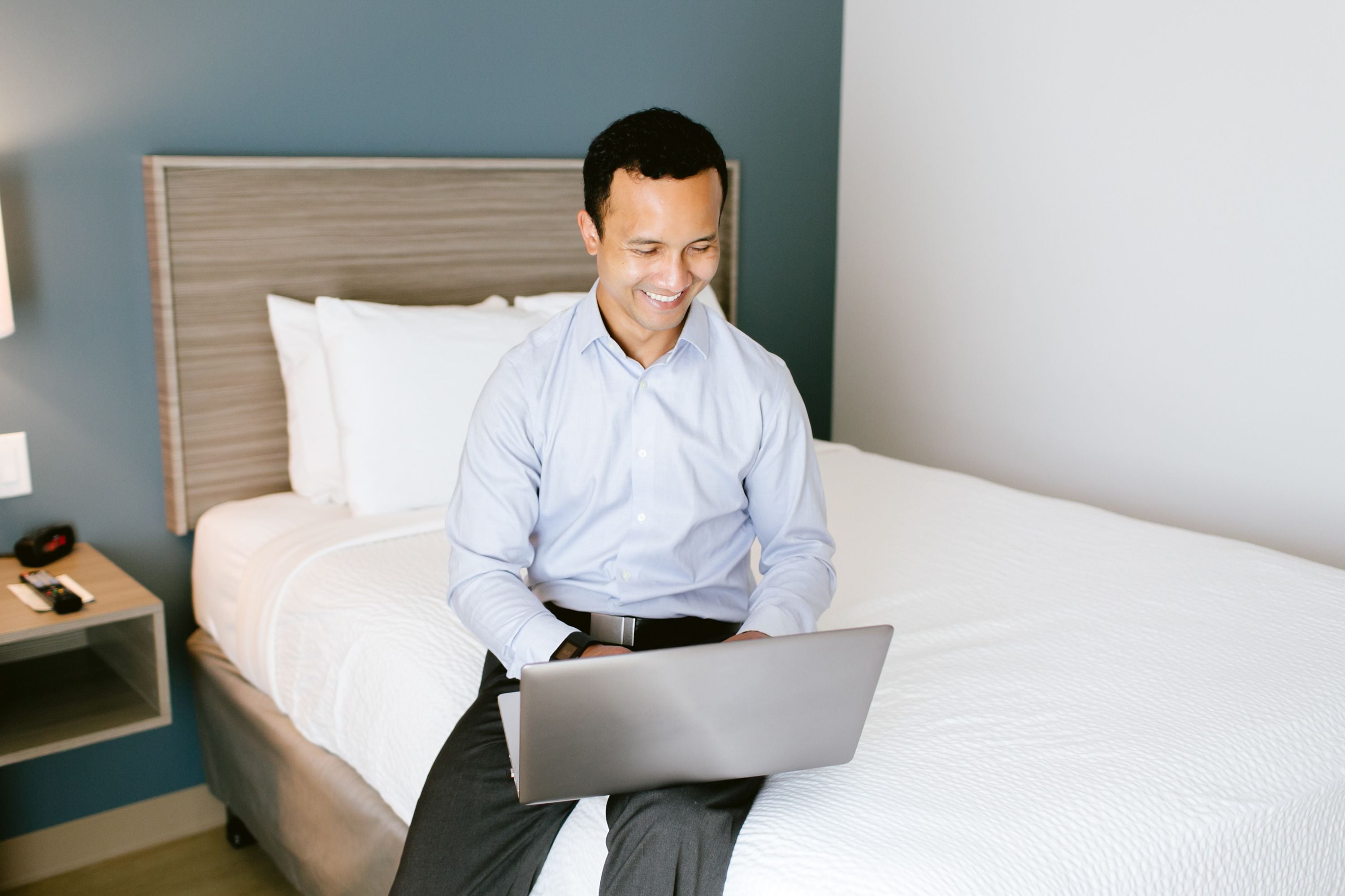 A person sitting on a bed with a laptop, a bedside table with a phone, and a nightstand with a lamp.