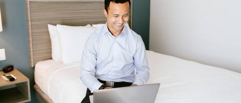 A person sitting on a bed with a laptop, a bedside table with a phone, and a nightstand with a lamp.