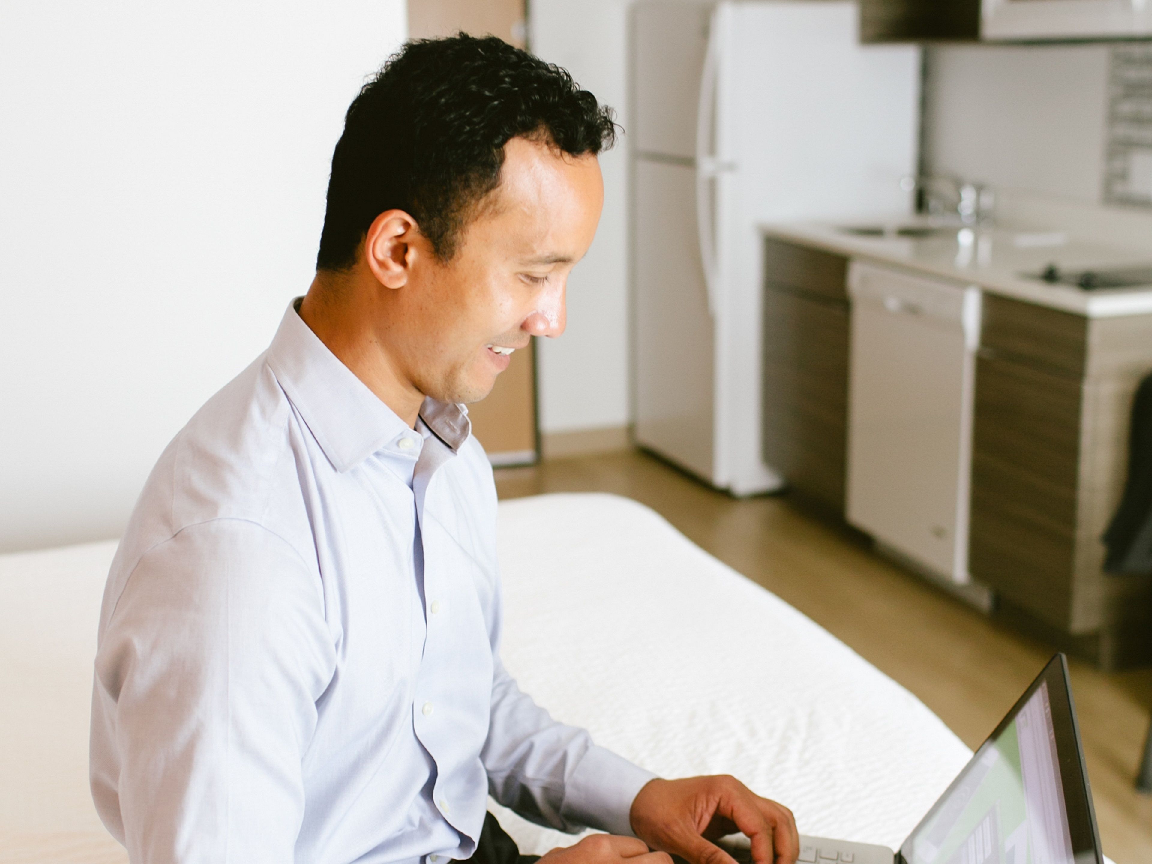 A man in a light blue shirt using a laptop in a modern hotel room with a bed, a small kitchenette, and a white wall.