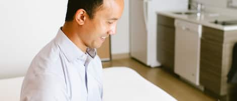 A man in a light blue shirt using a laptop in a modern hotel room with a bed, a small kitchenette, and a white wall.