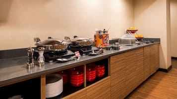 A buffet setup with various food containers, a coffee machine, and a fruit display.