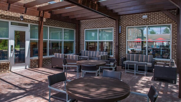 Ein Außenbereich mit einer Terrasse, Ziegelmauern, großen Fenstern und verschiedenen Sitzgelegenheiten, darunter runde Tische und Stühle.