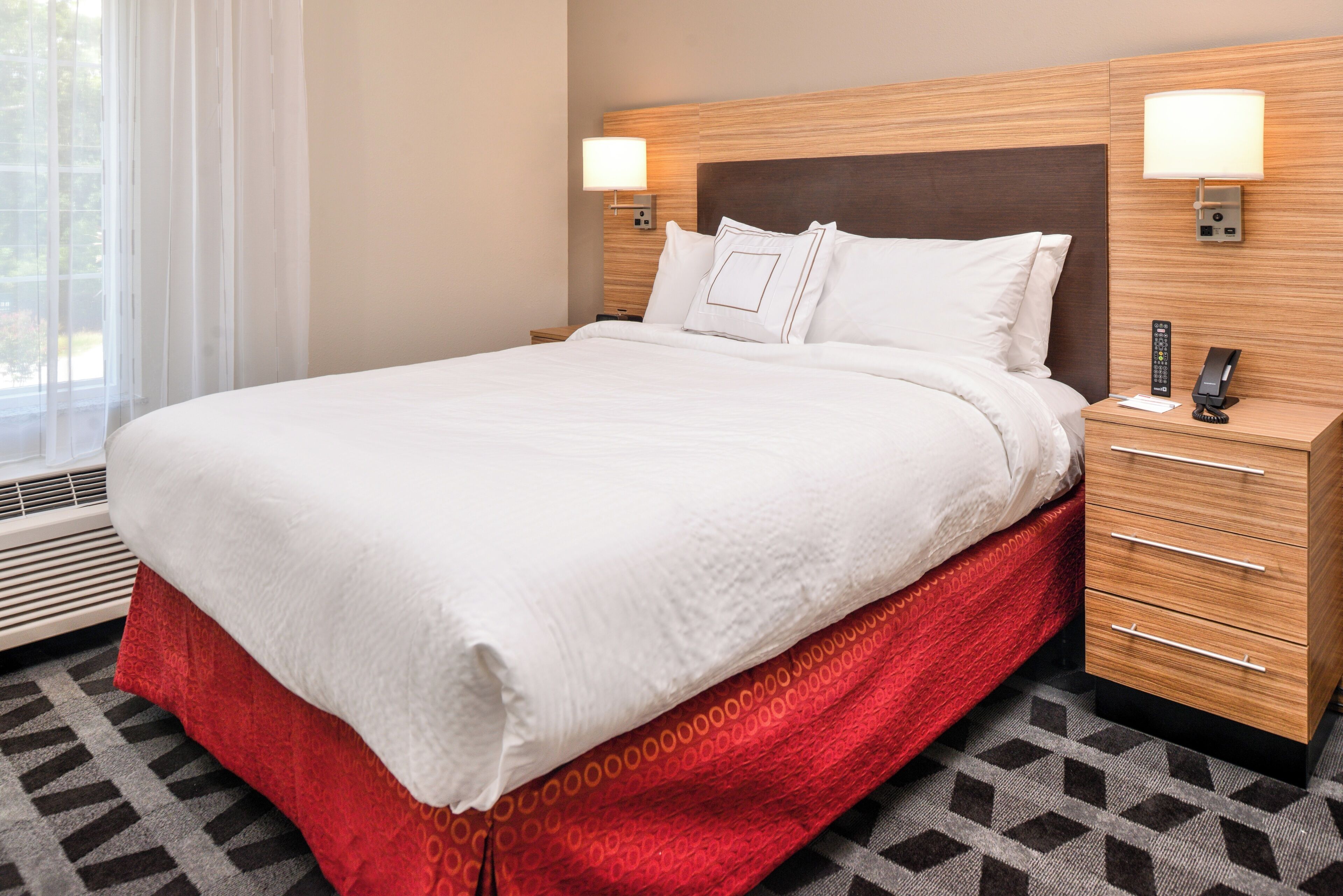 Une chambre d’hôtel avec un lit, une table de chevet avec une lampe, un téléphone et une fenêtre avec des rideaux.