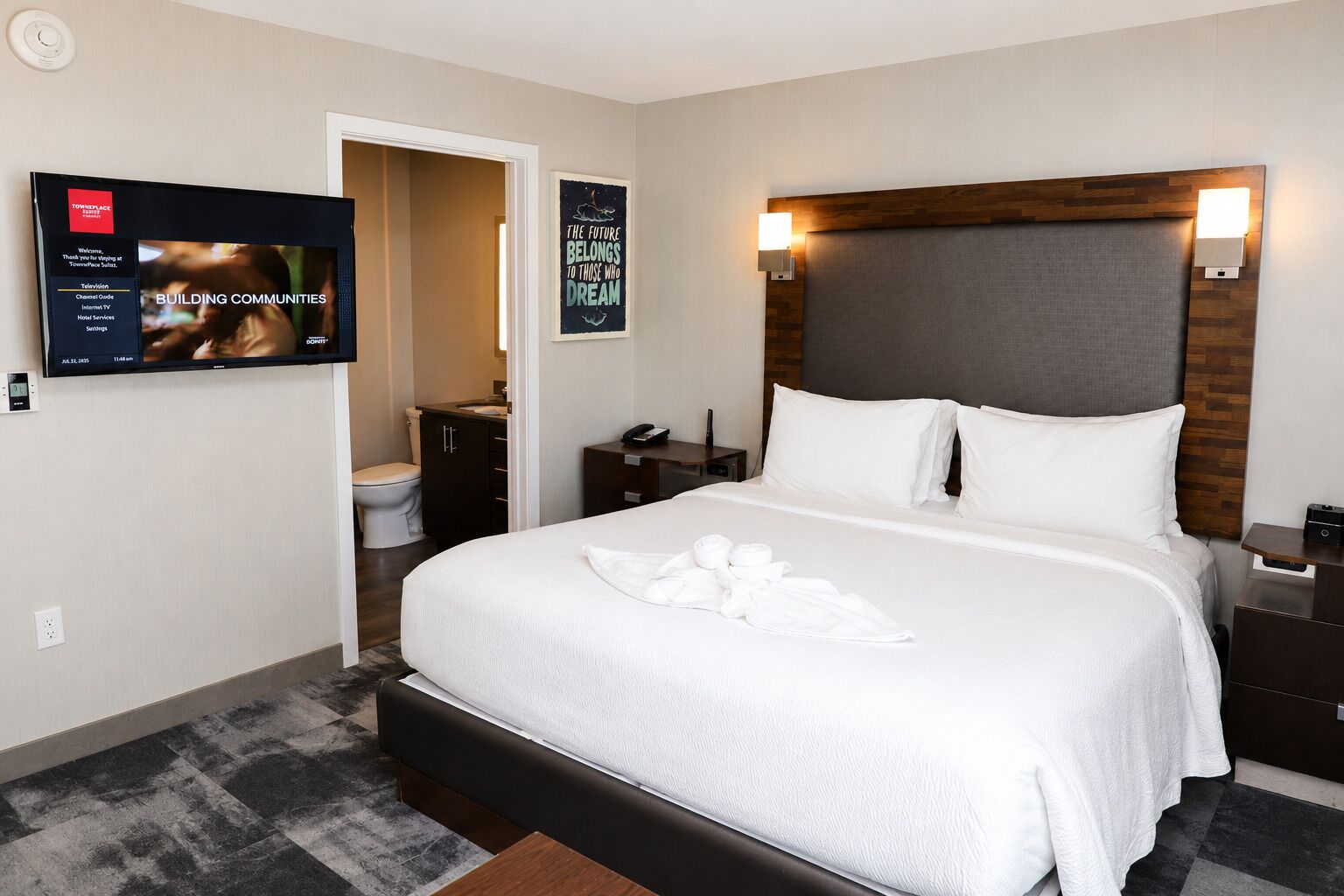 A hotel room with a large bed, a television mounted on the wall, a wooden headboard, and a poster on the wall.