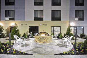A courtyard with a stone fire pit surrounded by white chairs, a building with multiple windows in the background, and a well-maintained garden with flowers and shrubs.
