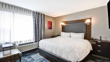 A hotel room with a large bed, a bedside table, a chair, a heater, a wall-mounted light, and a poster on the wall.