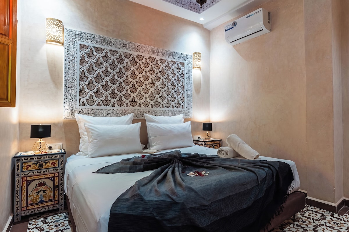 A hotel room with a bed, two bedside tables, a decorative headboard, a wall-mounted air conditioner, and a framed picture on the wall.