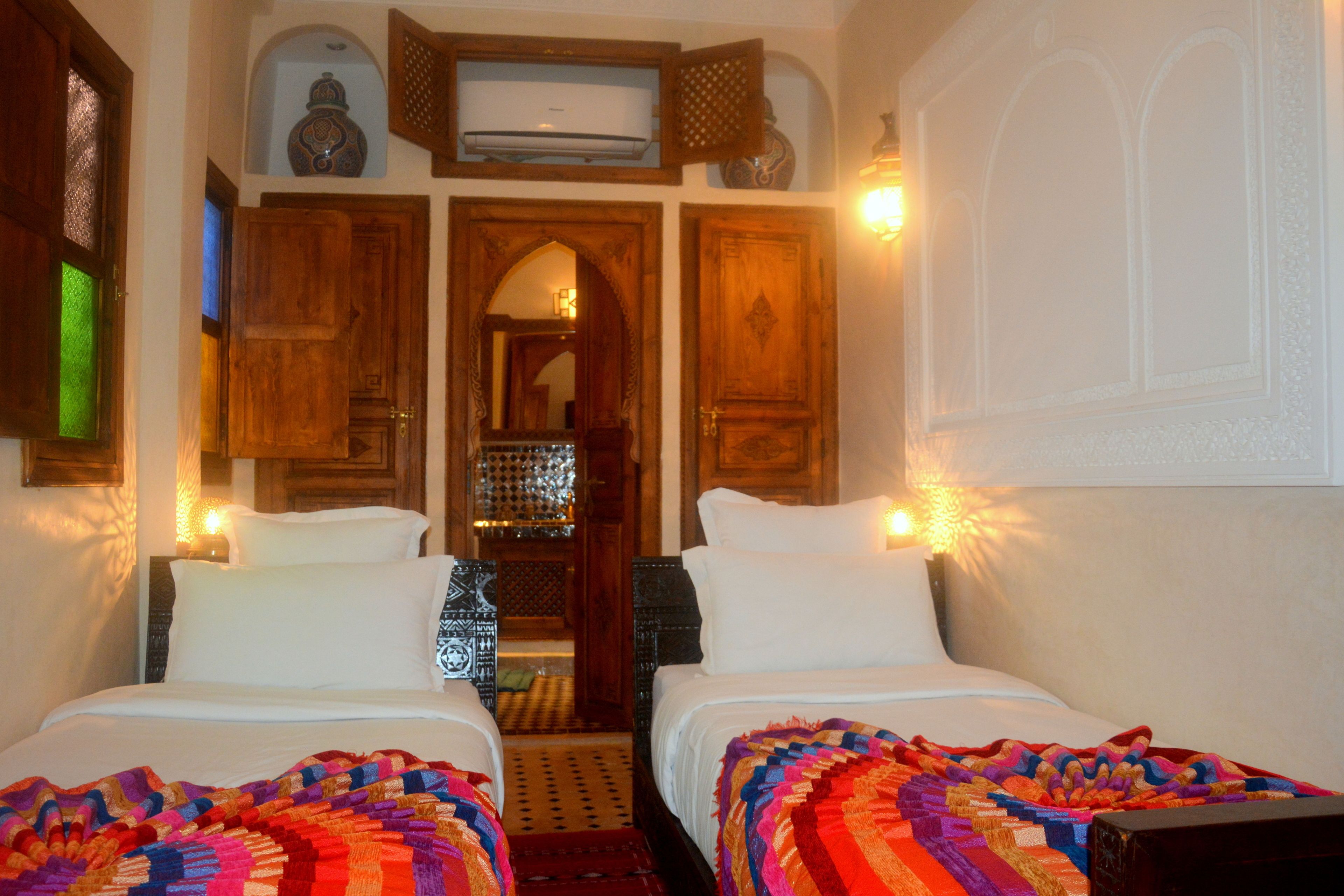 Quarto com duas camas, porta de madeira e um corredor pequeno que leva a um banheiro.