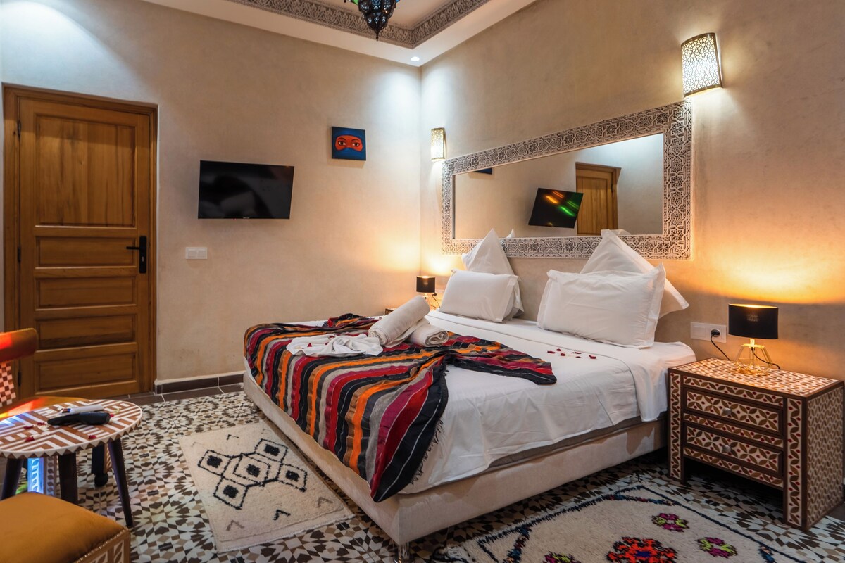 A hotel room with a bed, a small table, a chair, a TV, and a decorative mirror.