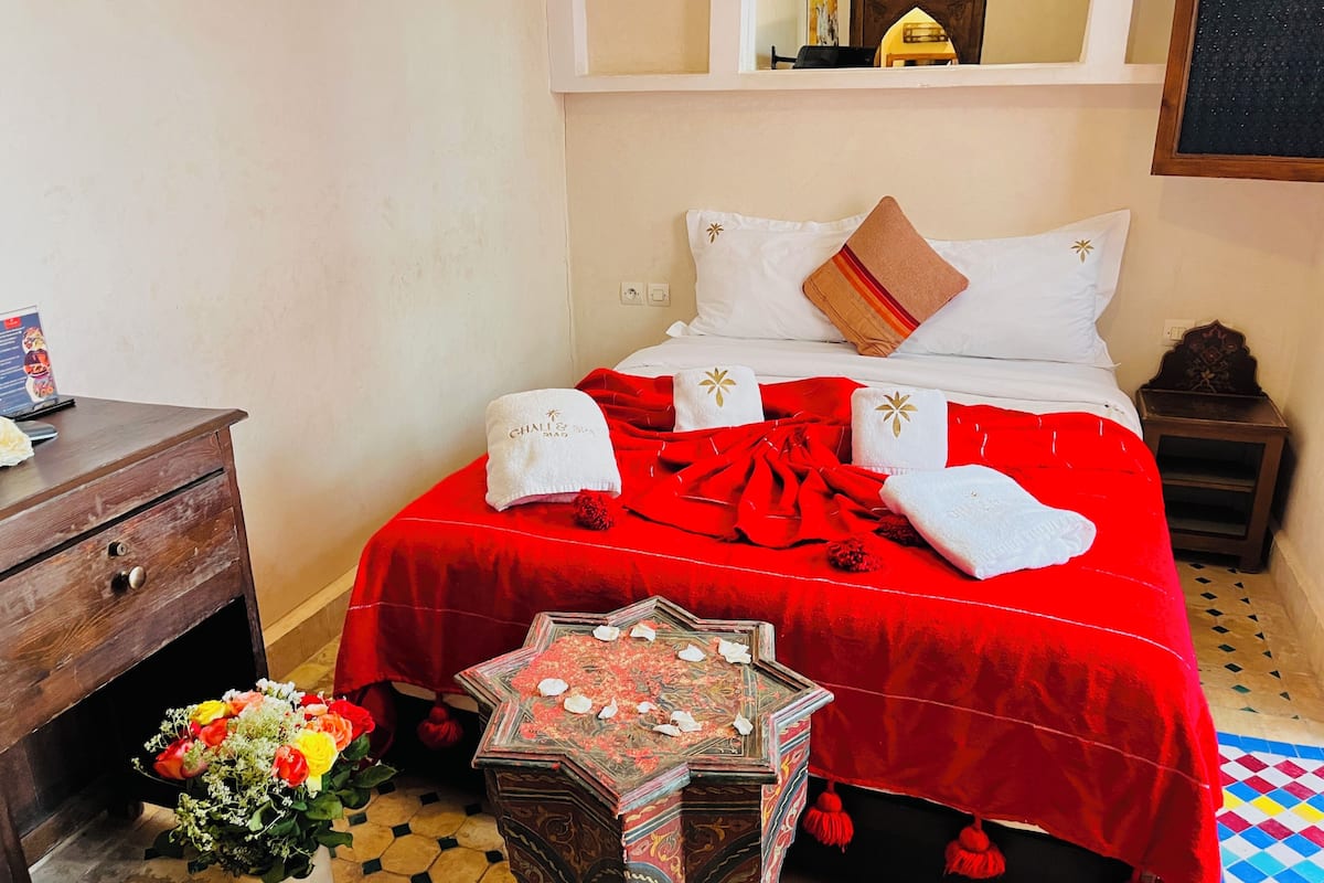 Een keurig ingerichte hotelkamer met een rode sprei, witte kussens met gouden details, een houten nachtkastje en een decoratietafel met bloemen.