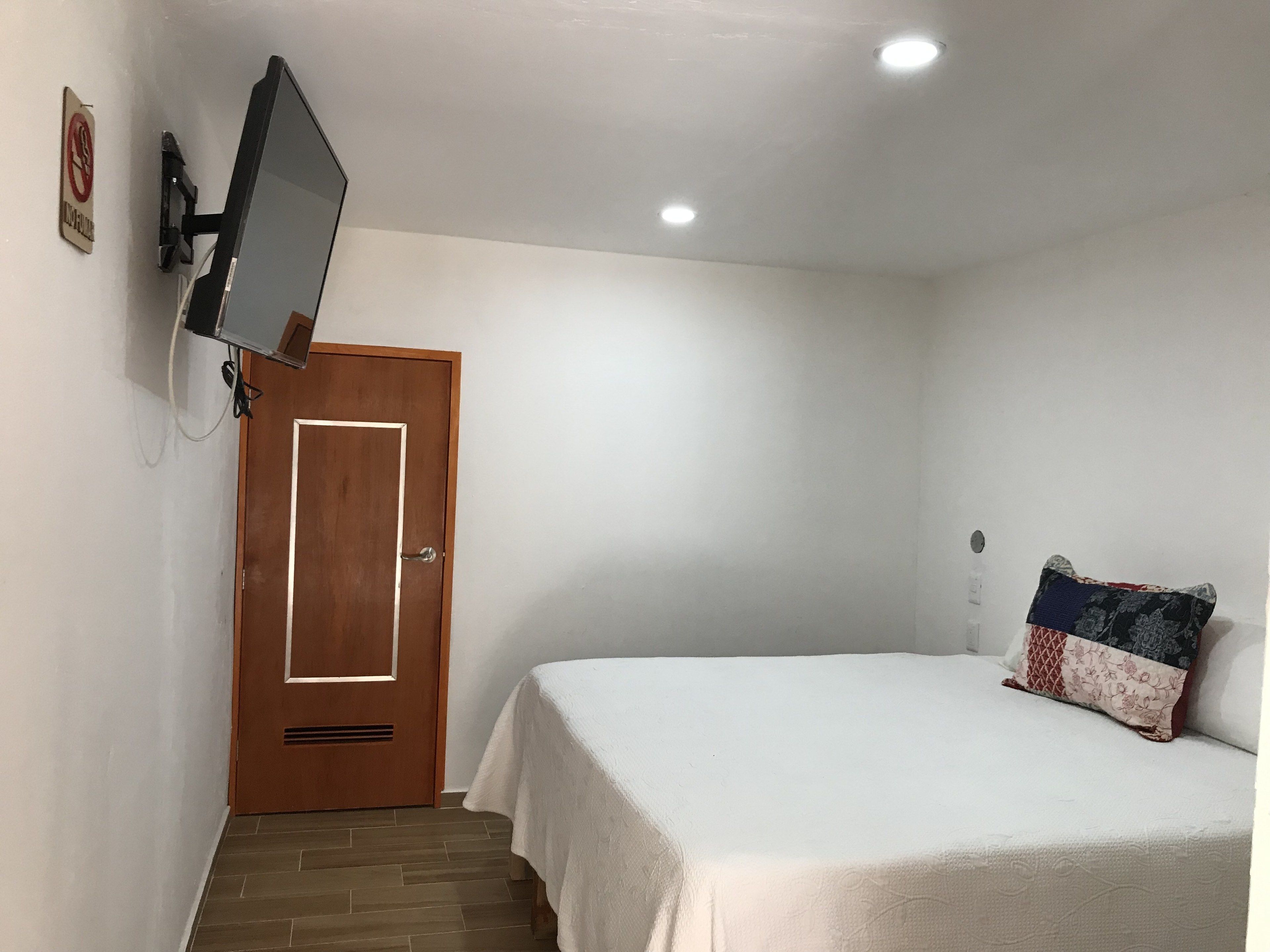 A bedroom with a bed, a wooden door, a mounted TV, and a small wall-mounted light fixture.