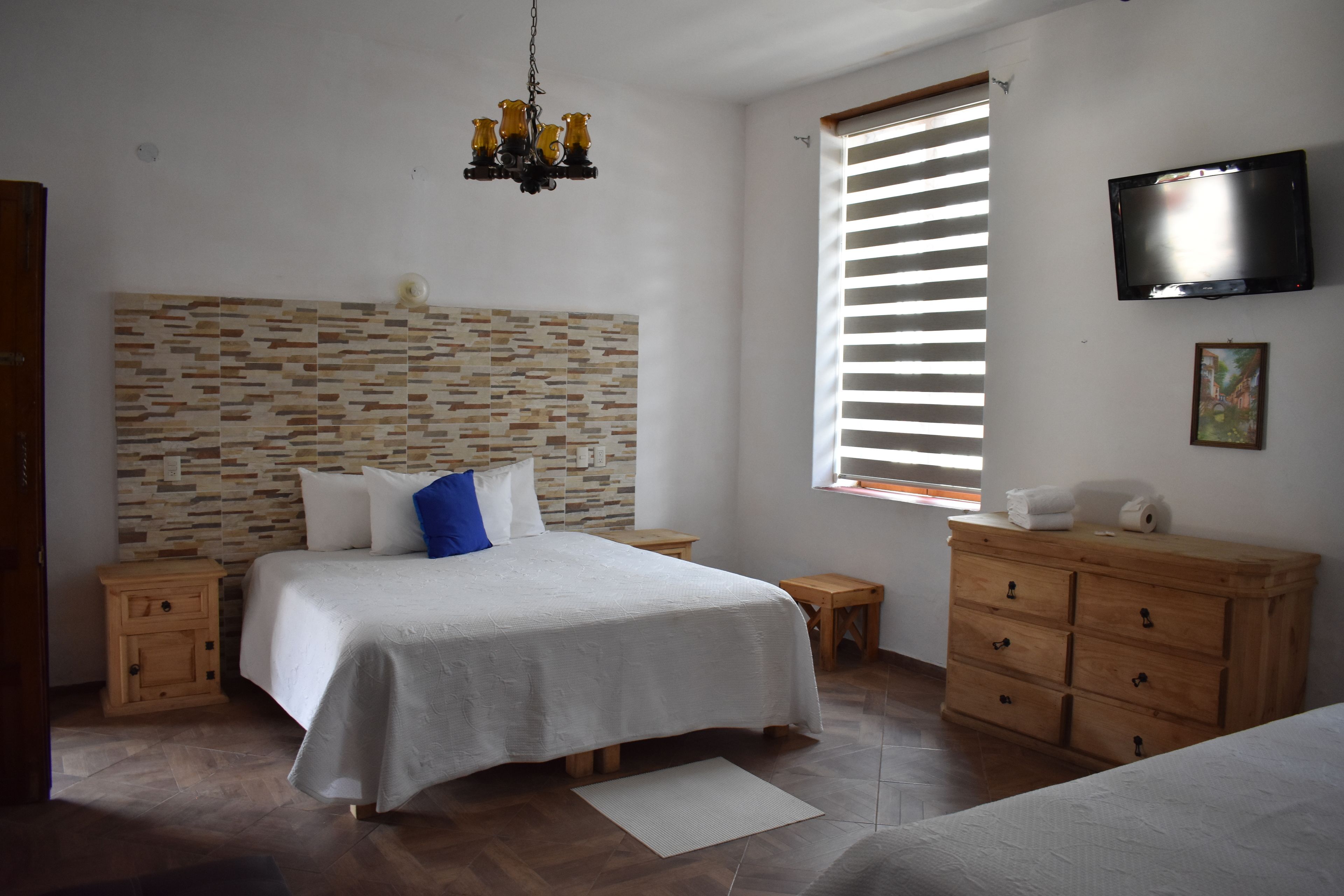 A bedroom with a bed, wooden nightstands, a dresser, a TV, and a window with blinds.