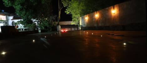 An outdoor area with a wet surface, possibly a courtyard, with trees and a wall with lights.