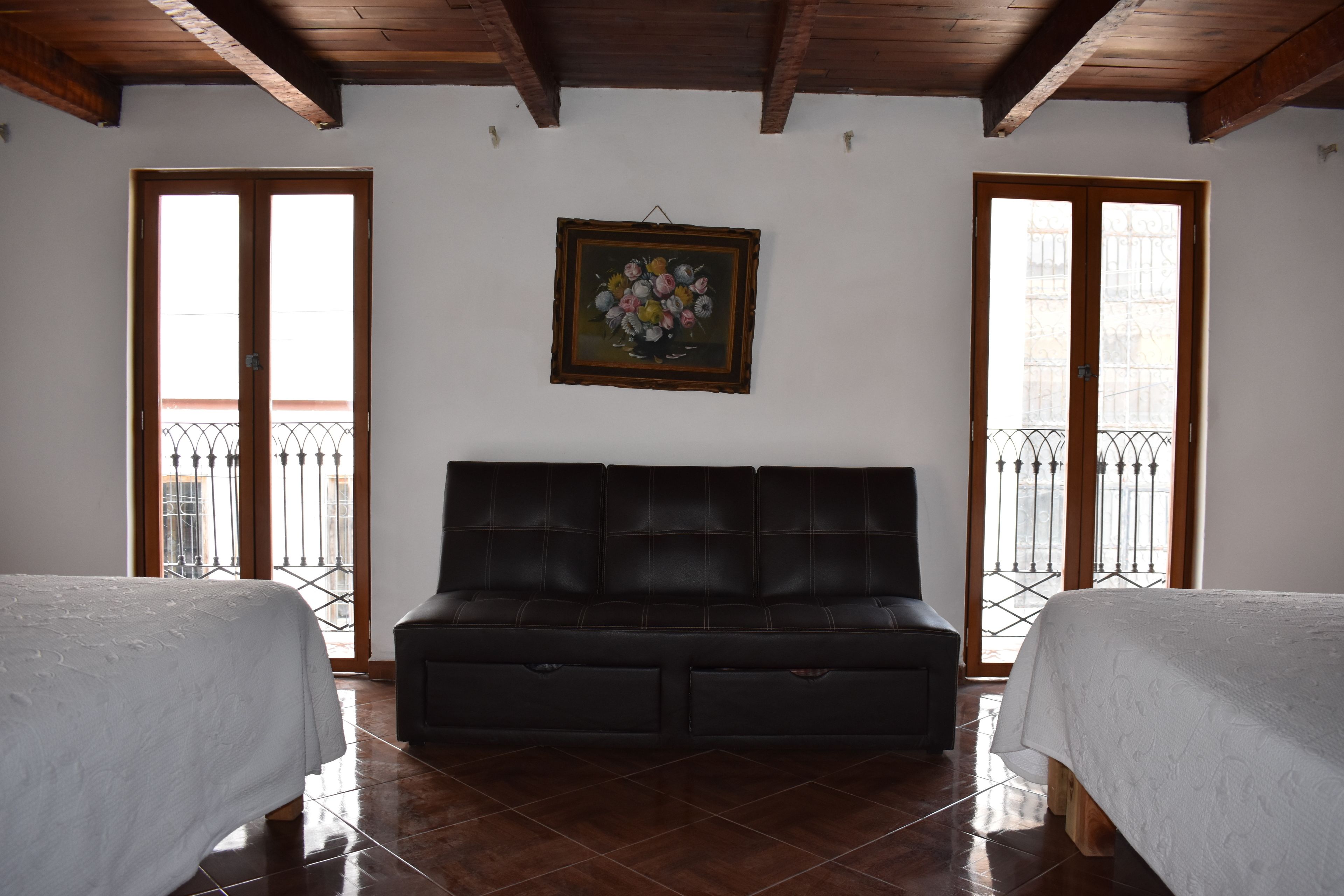 A room with a black leather sofa, two beds with white linens, and a framed floral painting on the wall.