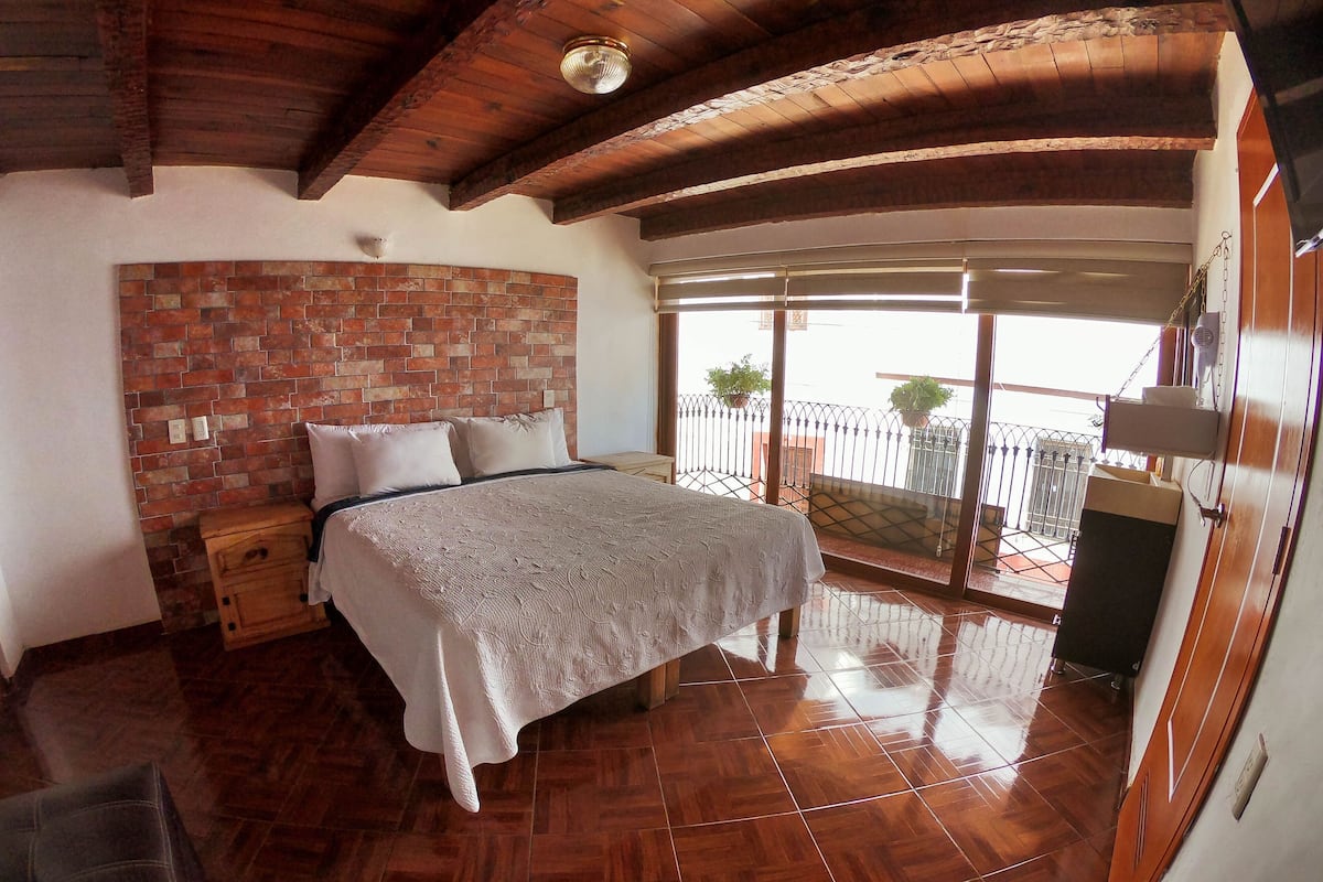 Un dormitorio con pared de ladrillo decorativa, cama con cubrecama blanco, suelo de madera y balcón con vistas.