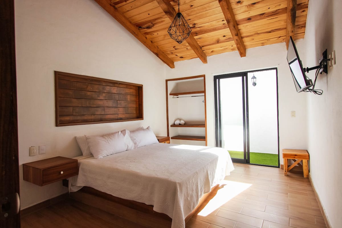 Un dormitorio con techo de madera, una cama con colcha blanca, una mesita de noche, un banco de madera y un ventanal corredero que da a un balcón.
