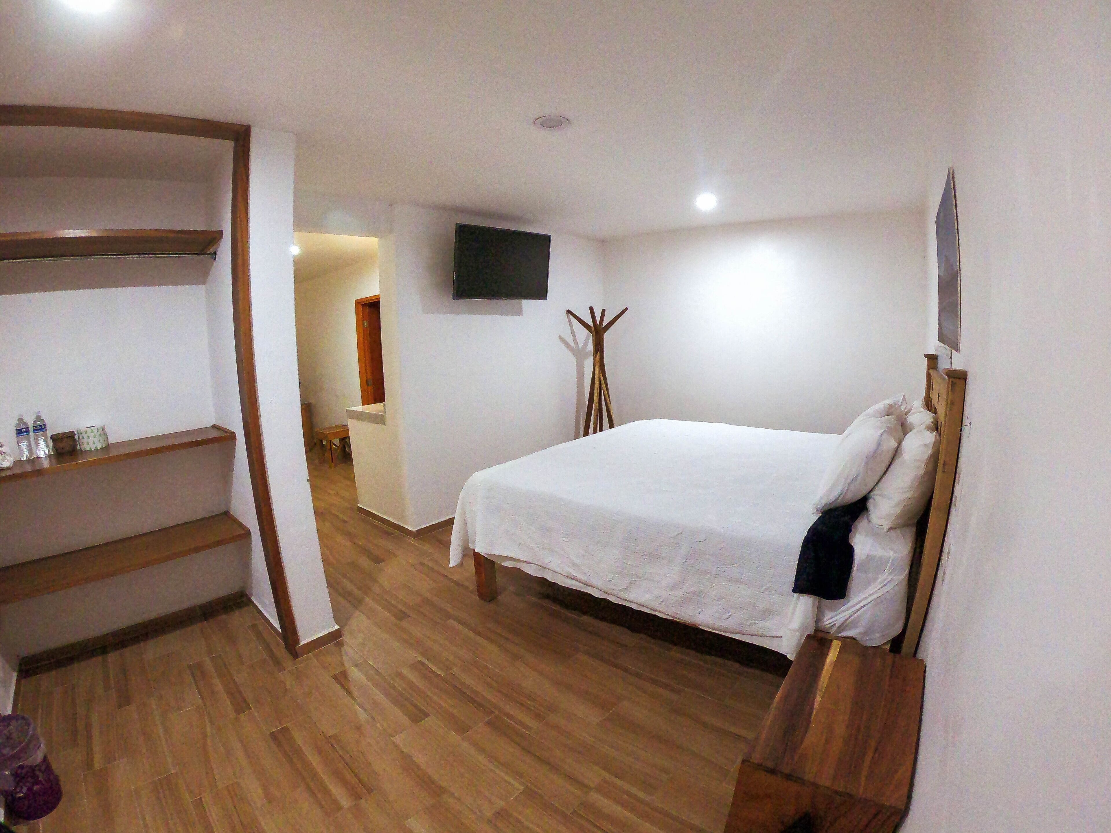 A bedroom with a wooden floor, a bed with white bedding, a TV mounted on the wall, and a wooden bench.