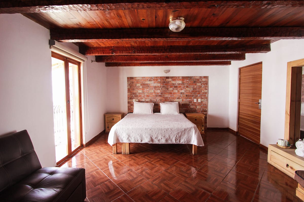 Un dormitorio con techo de madera, pared de ladrillo, cama con ropa blanca, mesita de noche de madera, cómoda de madera y una silla.