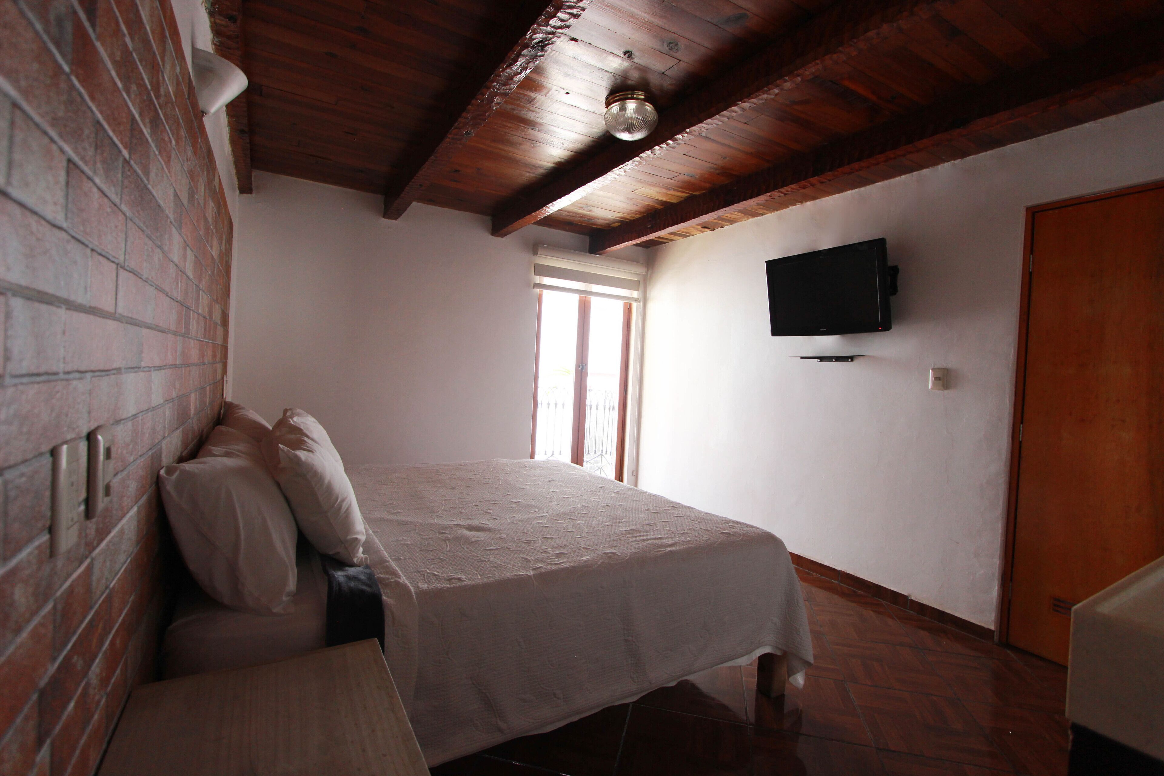 A bedroom with a bed, a brick wall, a wooden ceiling, a television, and a door.