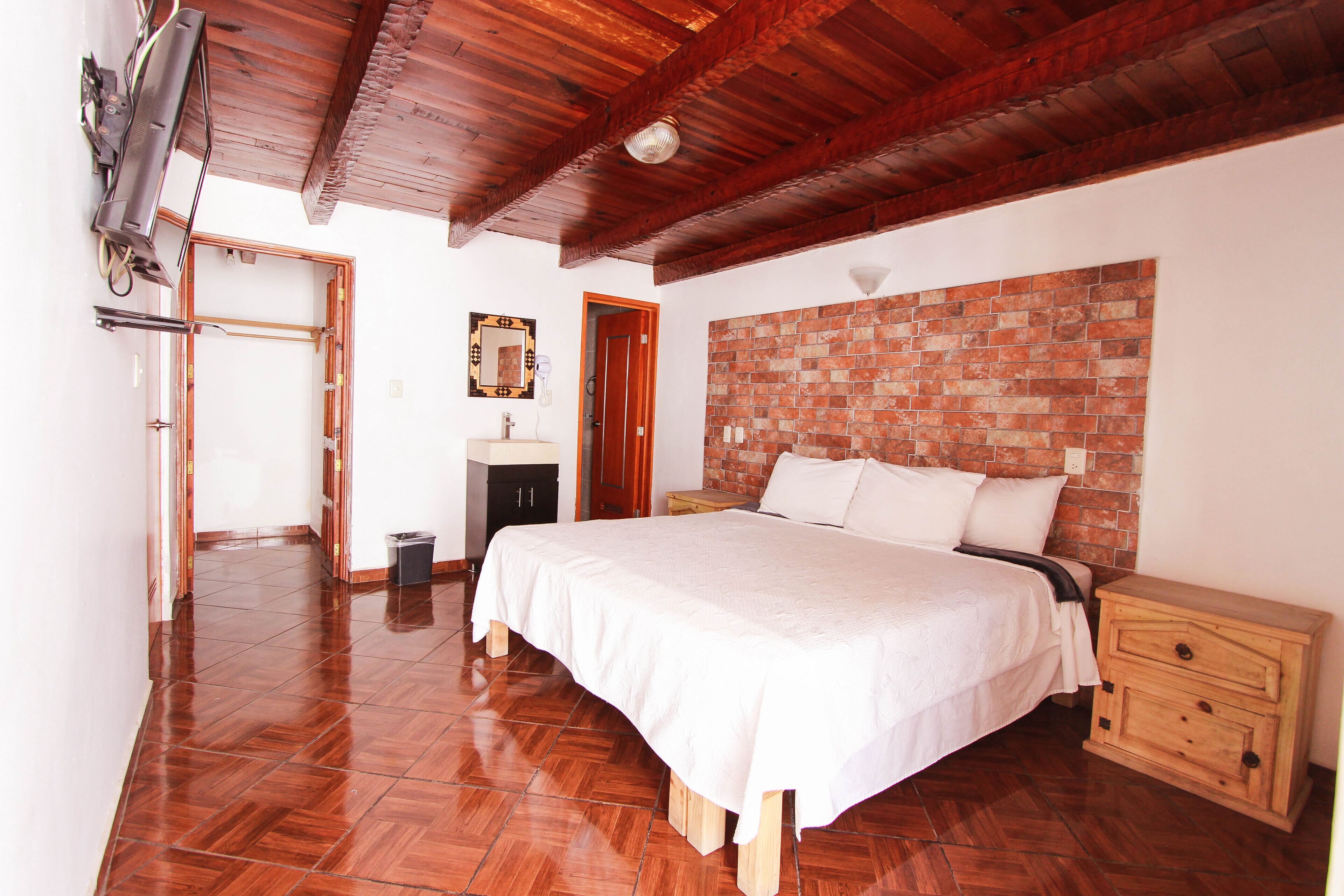 A bedroom with a wooden ceiling, a brick-patterned headboard, a large bed, a wooden nightstand, and a tiled floor.