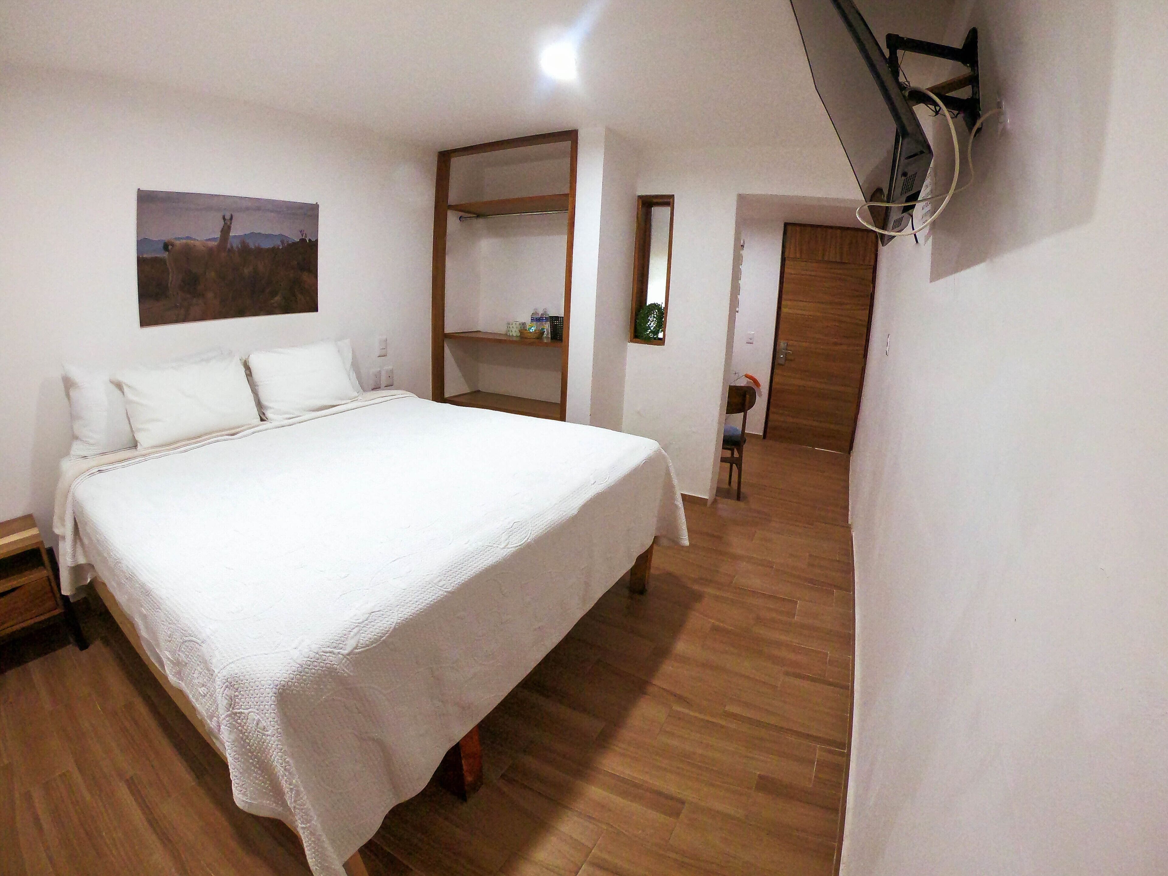 A bedroom with a large bed, a wooden floor, a wall-mounted shelf, and a door leading to another room.
