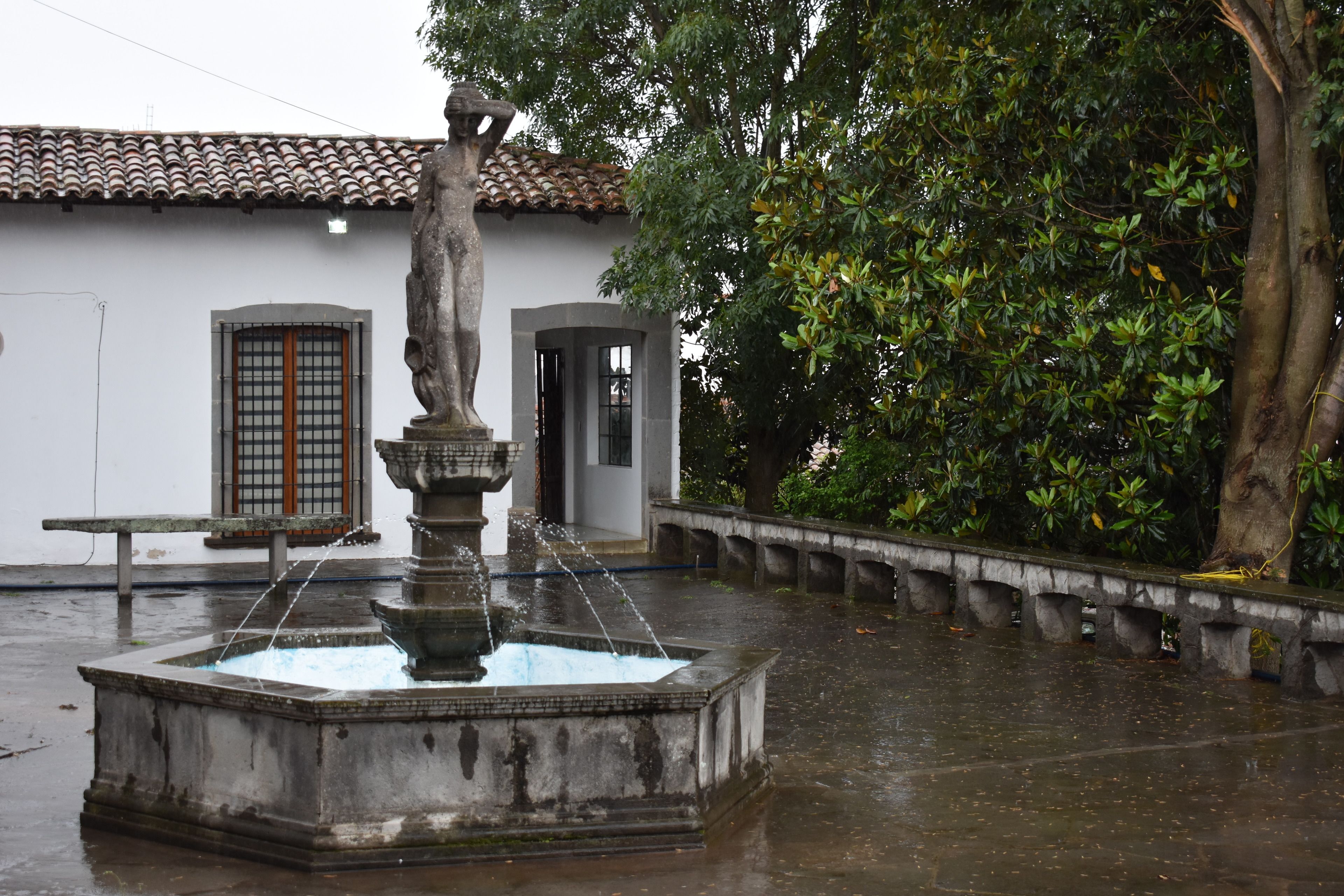 Una fuente de piedra con agua brotando de la parte superior, rodeada por una plaza húmeda y un edificio con techo de tejas al fondo.