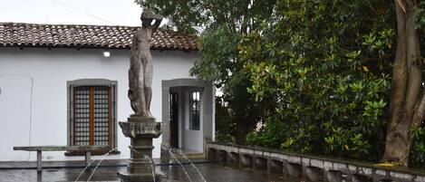 Una fuente de piedra con agua brotando de la parte superior, rodeada por una plaza húmeda y un edificio con techo de tejas al fondo.