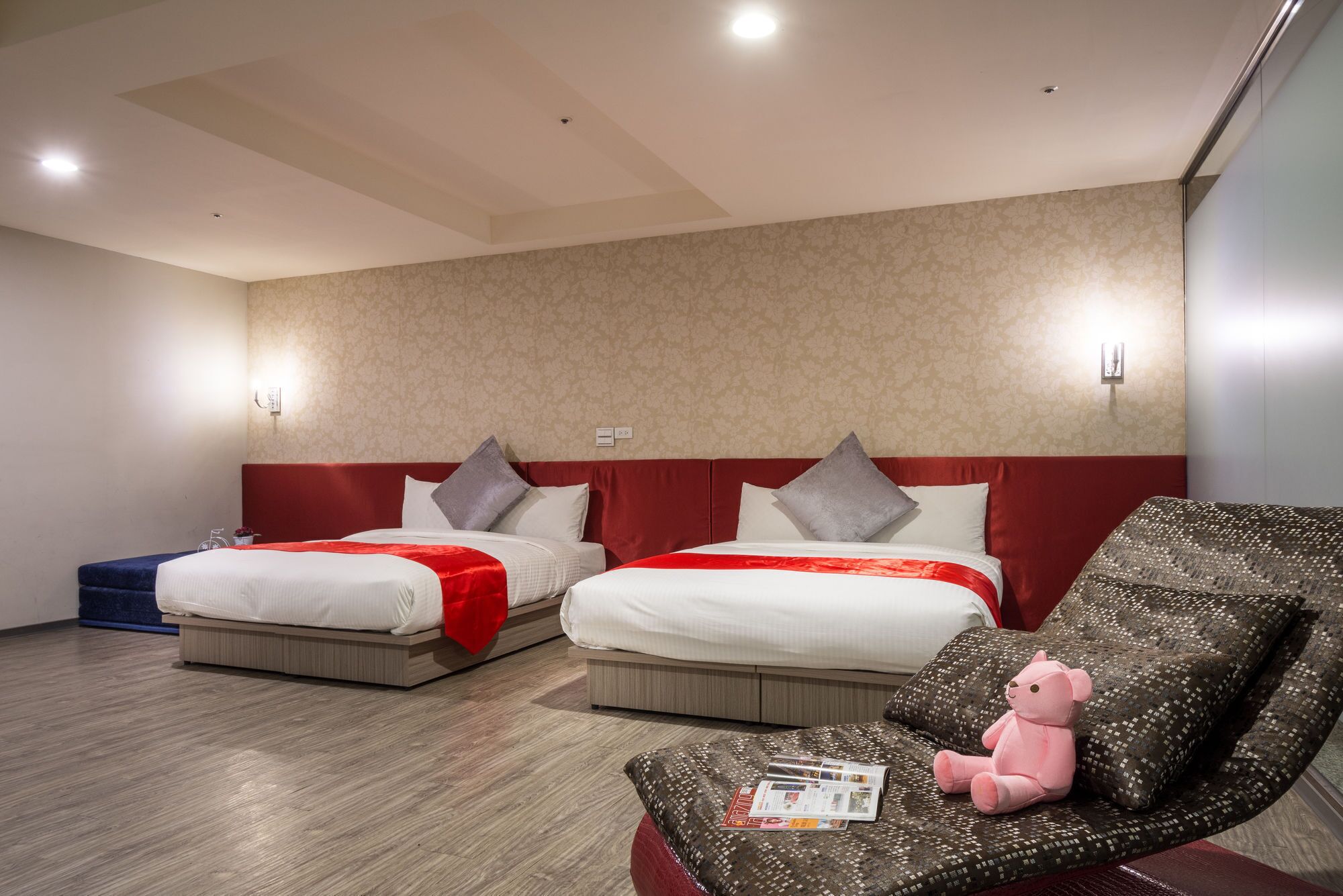 A hotel room with two beds, a red headboard, a wooden floor, and a seating area with a magazine and a pink teddy bear.