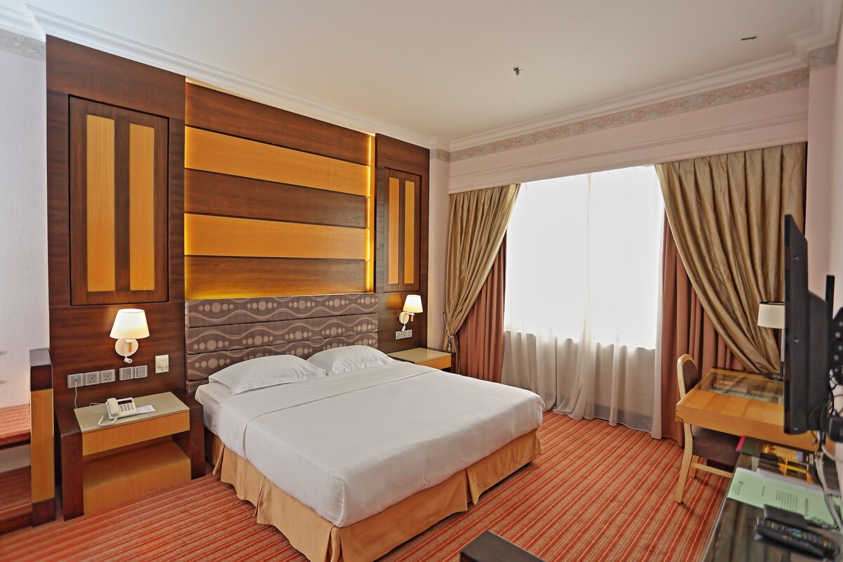 Una habitación de hotel con una cama grande, un escritorio con silla, televisión y ventana con cortinas.