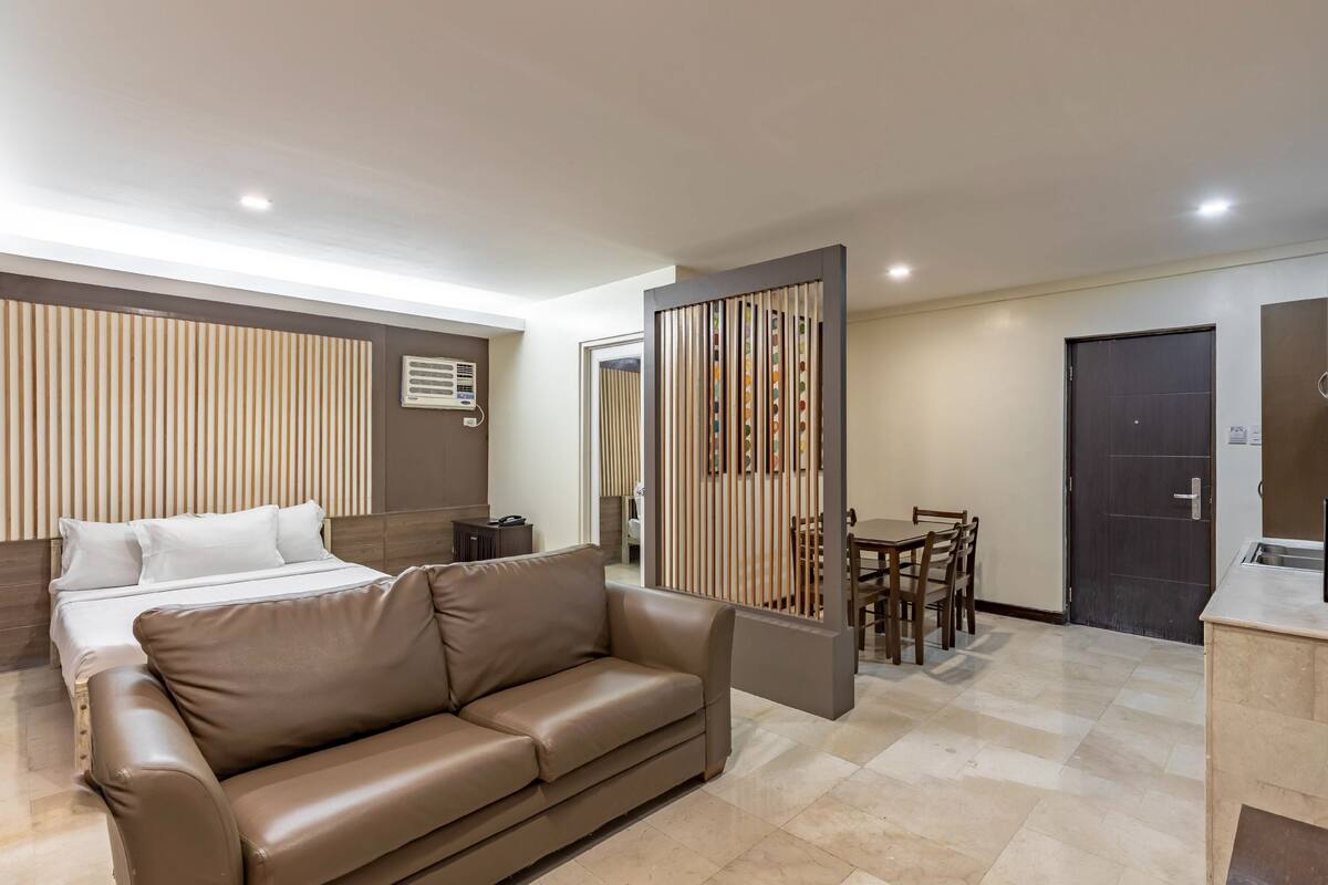 A modern hotel room with a bed, a leather sofa, a dining area, and a kitchen.