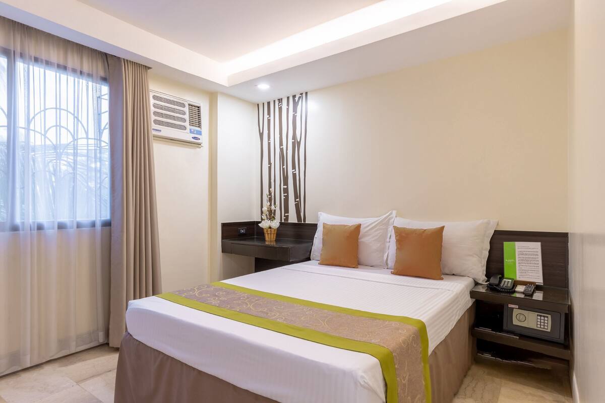 A hotel room with a bed, bedside table, and a nightstand with a phone and a safe.