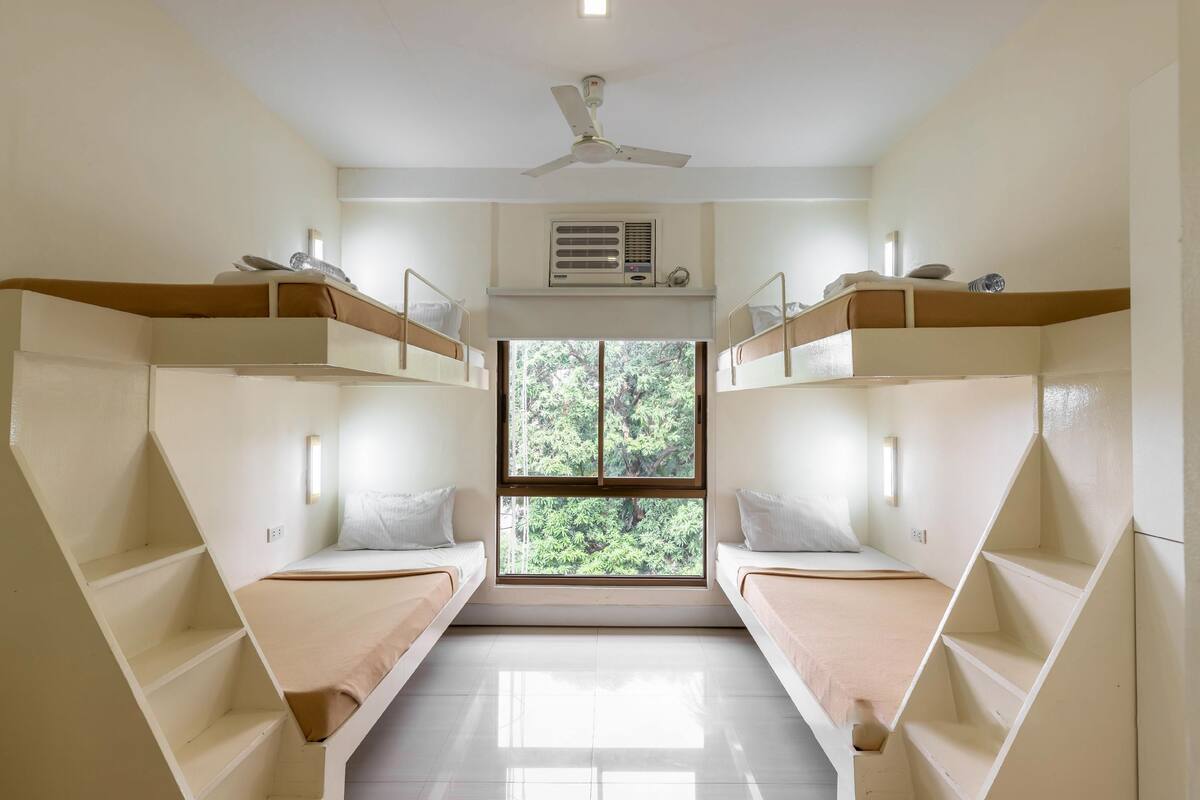A dormitory room with bunk beds, a ceiling fan, an air conditioning unit, and a window with a view of trees.