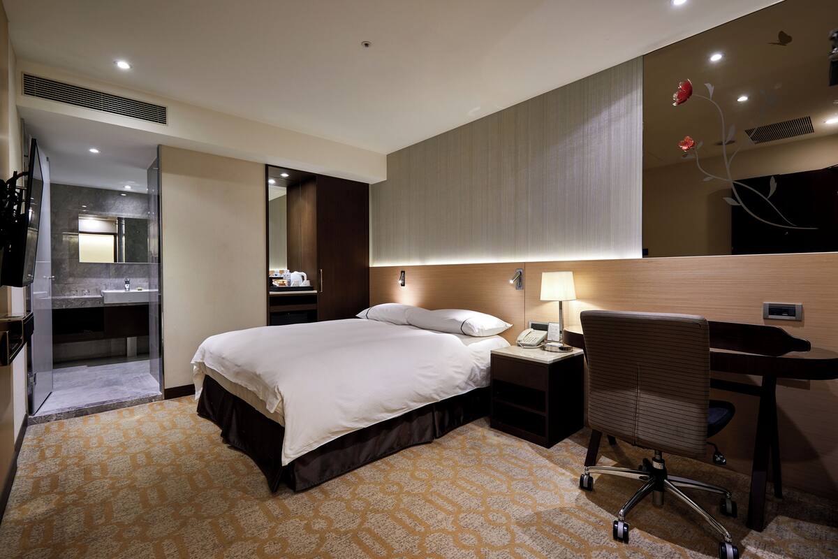 A hotel room with a bed, bedside table, desk, chair, and a bathroom with a sink and mirror.