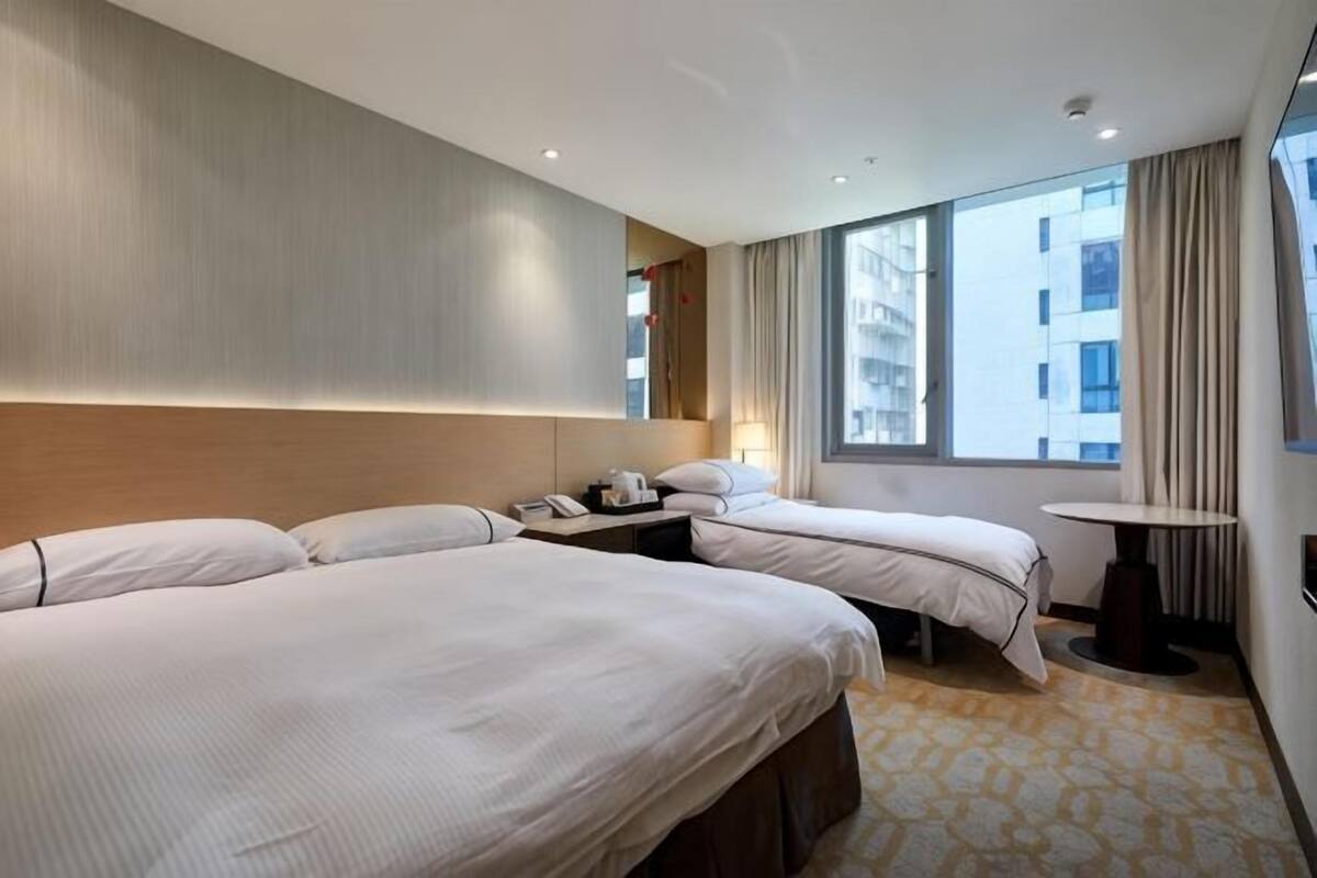 A hotel room with two beds, a small table, a nightstand, and a window with curtains.