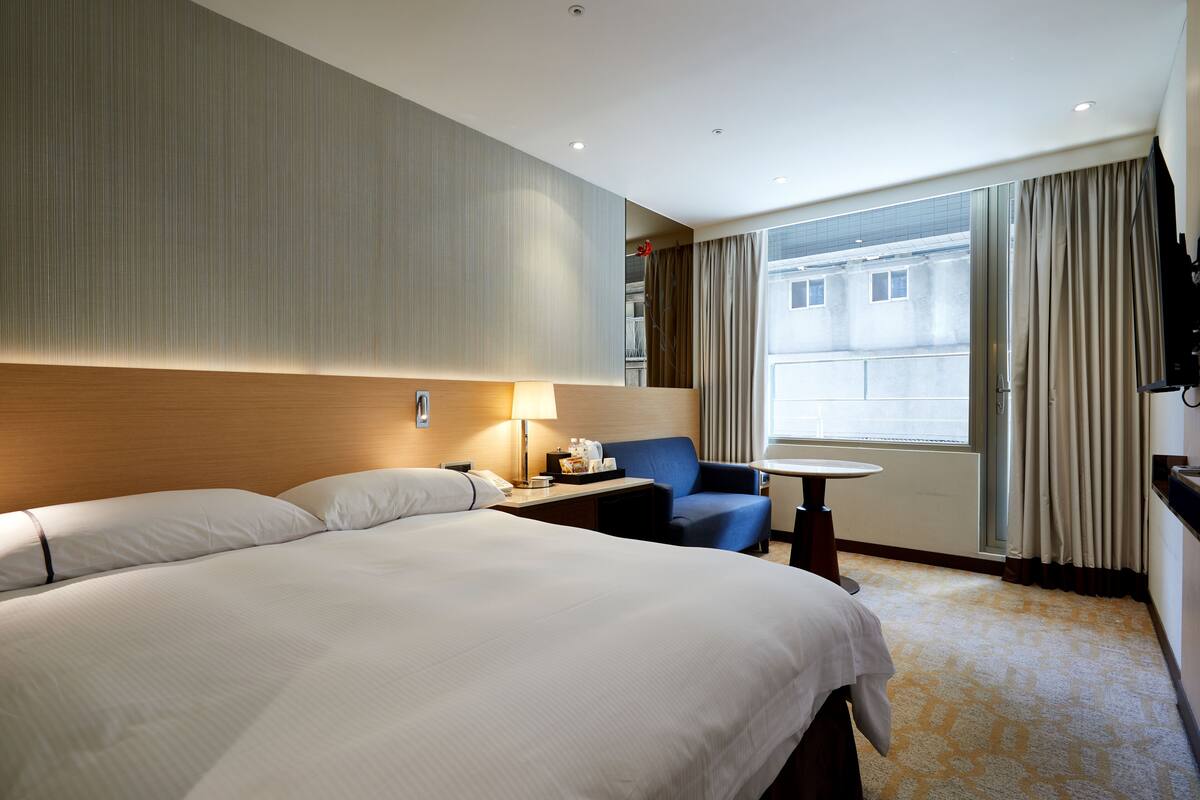 A hotel room with a large bed, a desk with a lamp, a chair, and a view of the city through the window.