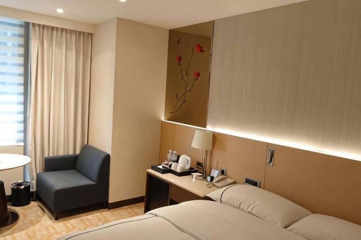 A hotel room with a bed, a desk with a lamp, a chair, a small table, and a window with curtains.