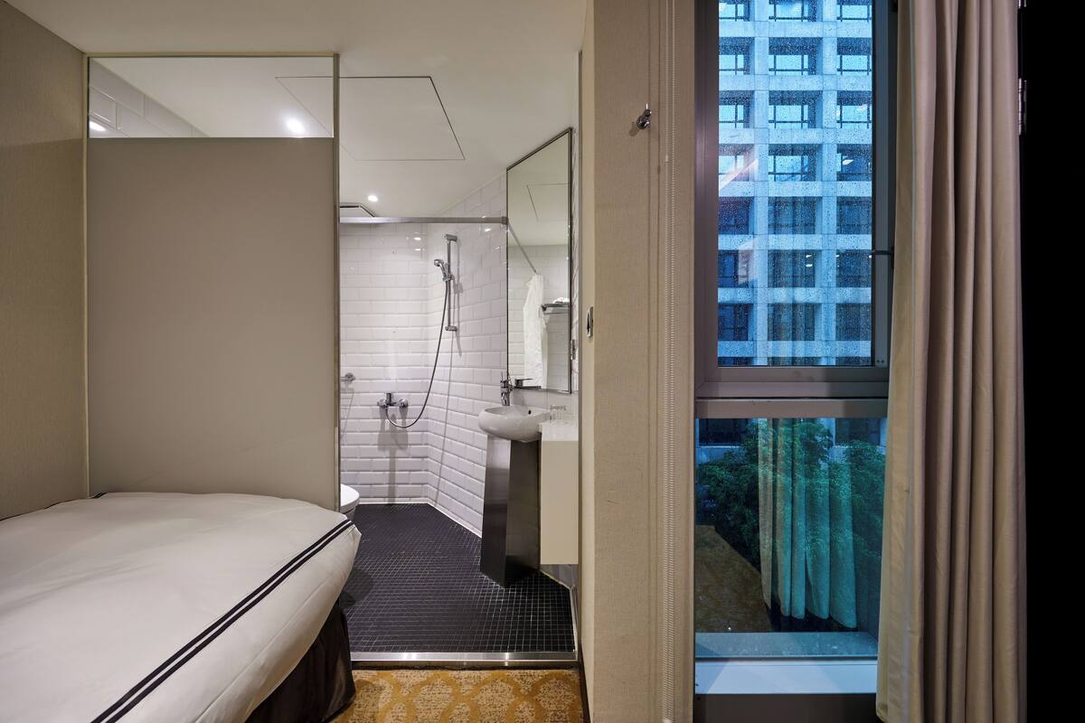 A modern hotel room with a bed, a bathroom with a shower, a sink, and a large window with curtains.