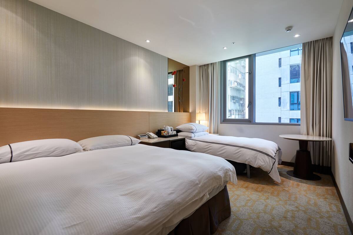 A hotel room with two beds, a small table, a nightstand, and a window with curtains.