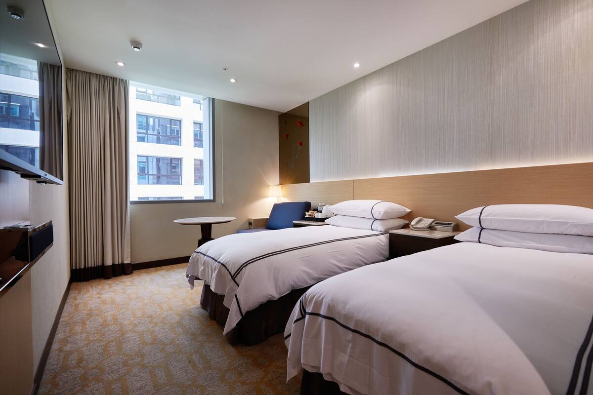 A hotel room with two beds, a small table, a chair, a lamp, and a window with curtains.