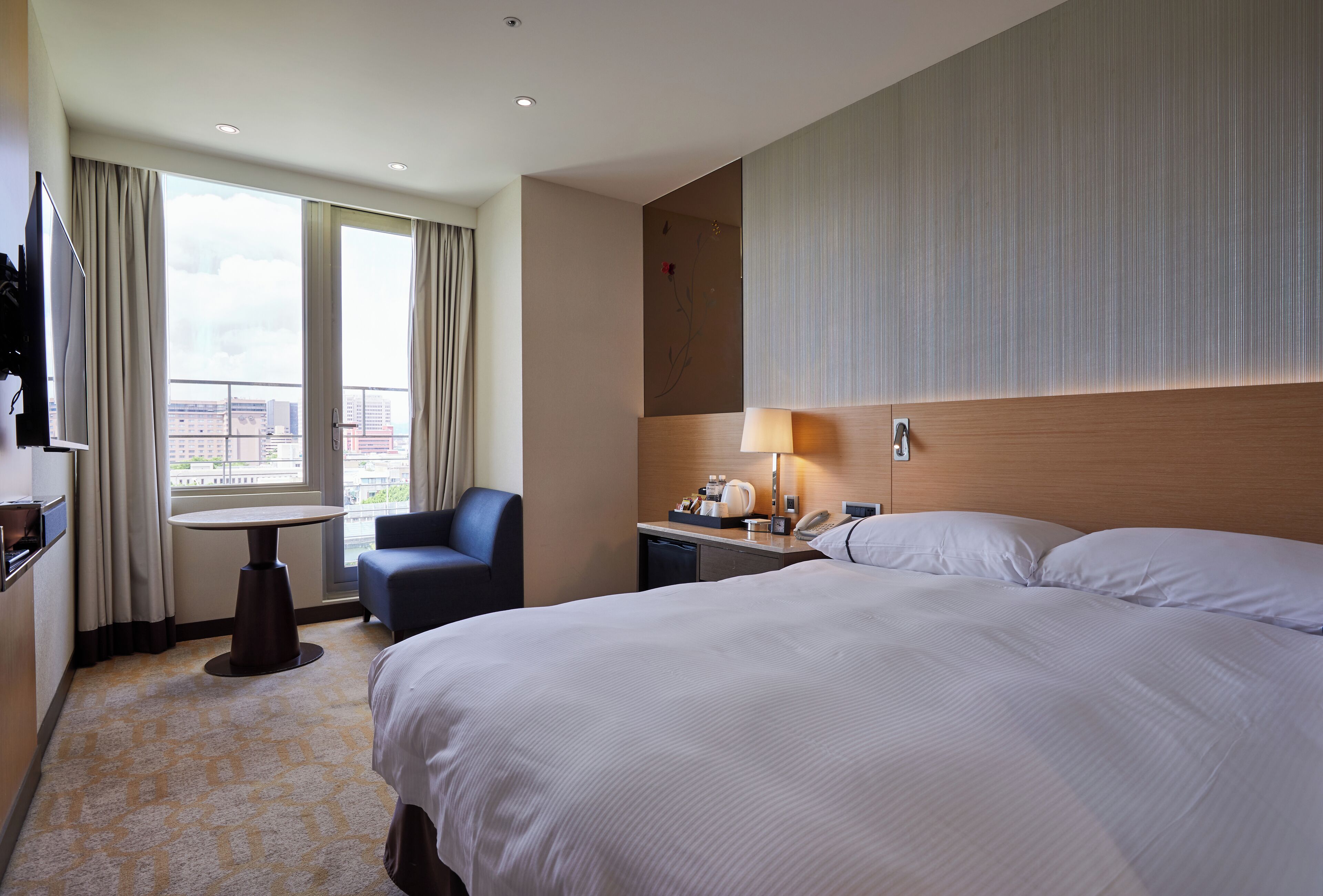 A hotel room with a large bed, a desk with a lamp, a chair, and a view of the city through large windows.