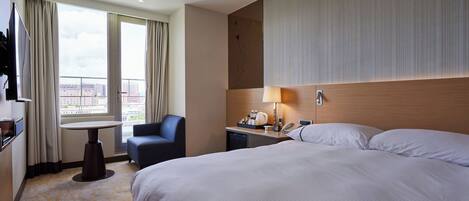 A hotel room with a large bed, a desk with a lamp, a chair, and a view of the city through large windows.