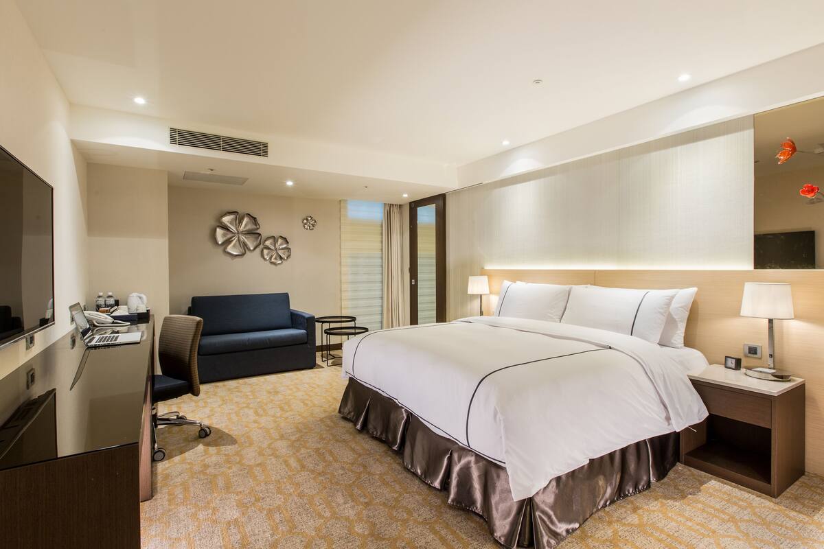 A hotel room with a large bed, a desk with a laptop, a chair, a sofa, and a TV.