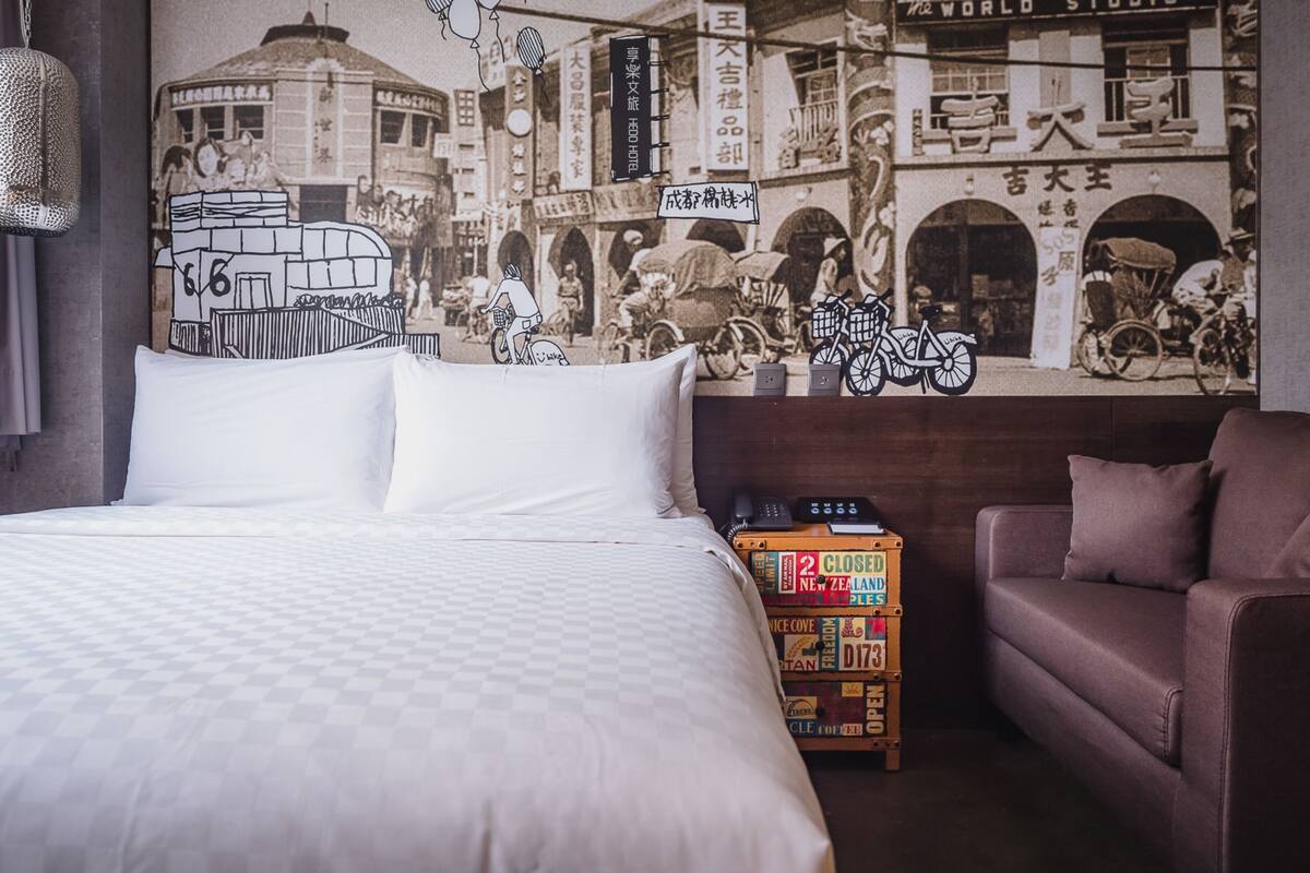Quarto de hotel com uma cama grande, um sofá, um mural na parede retratando uma cena de rua e uma pilha de malas.